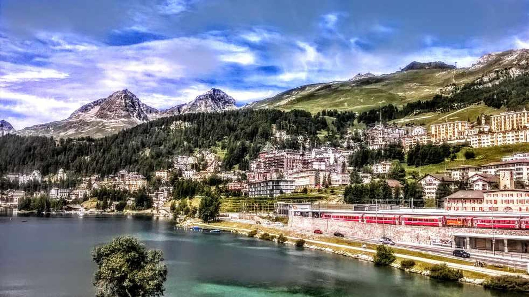 Scenic view of a lakeside town with a red train passing, surrounded by mountains and greenery