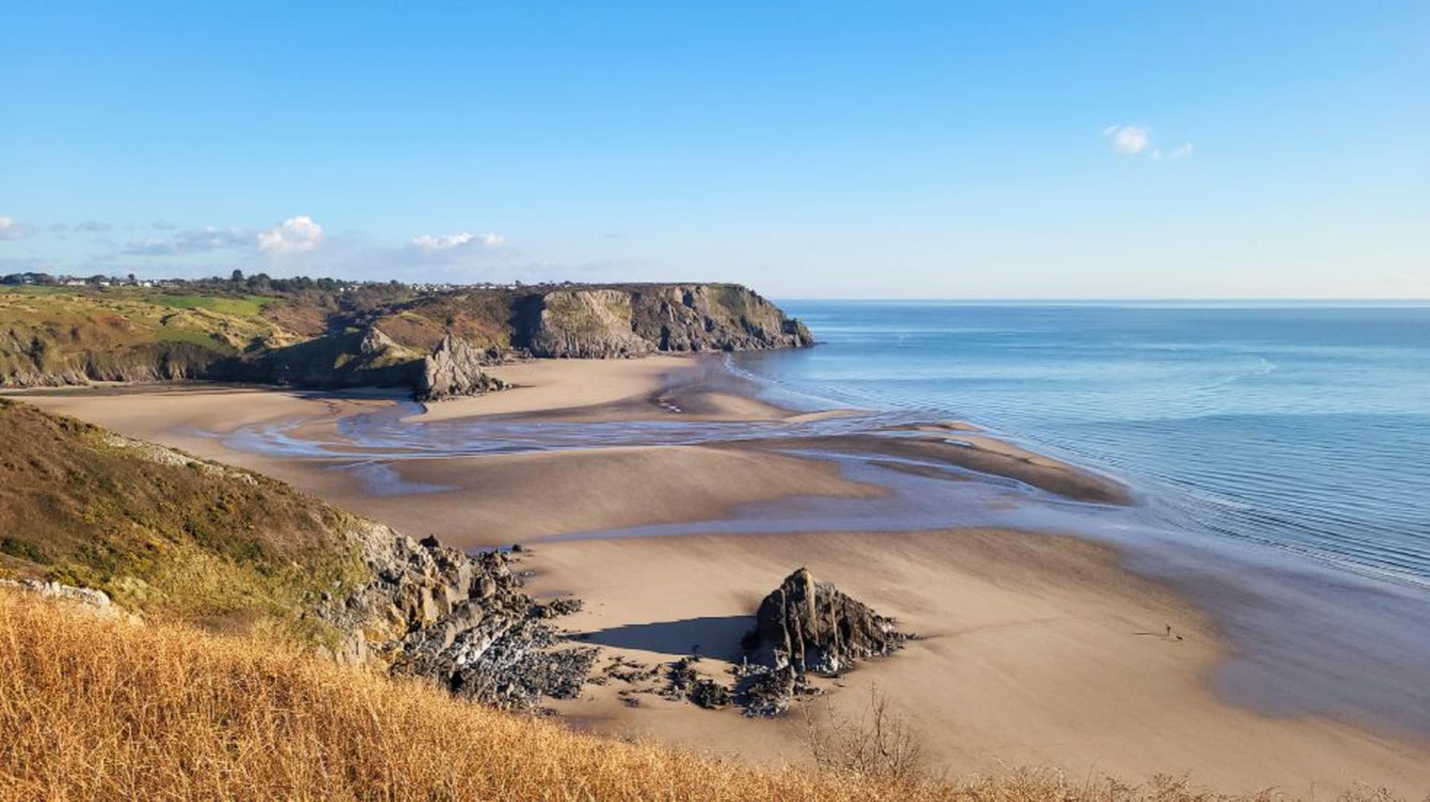 Coastal view of a sandy beach with rocky cliffs and clear blue skies. Gentle waves touch the shore, creating a serene seaside landscape.