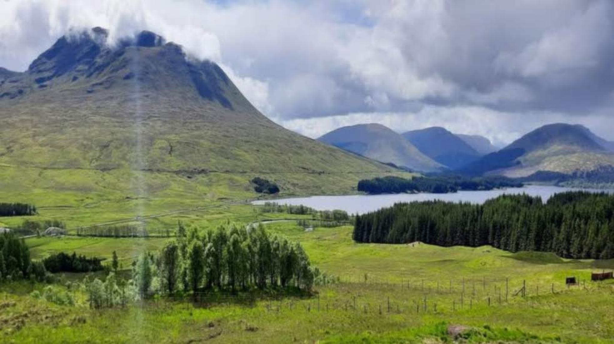 Scenic landscape with lush green fields, a tranquil lake, and mountains under a partly cloudy sky