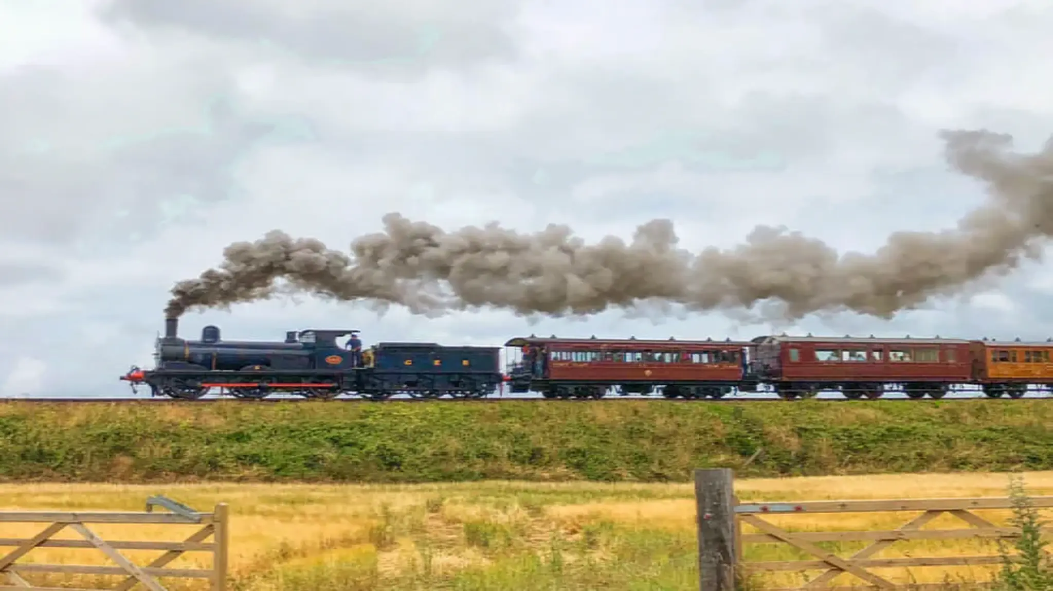 Steam train with billowing smoke travels along a track in a rural area