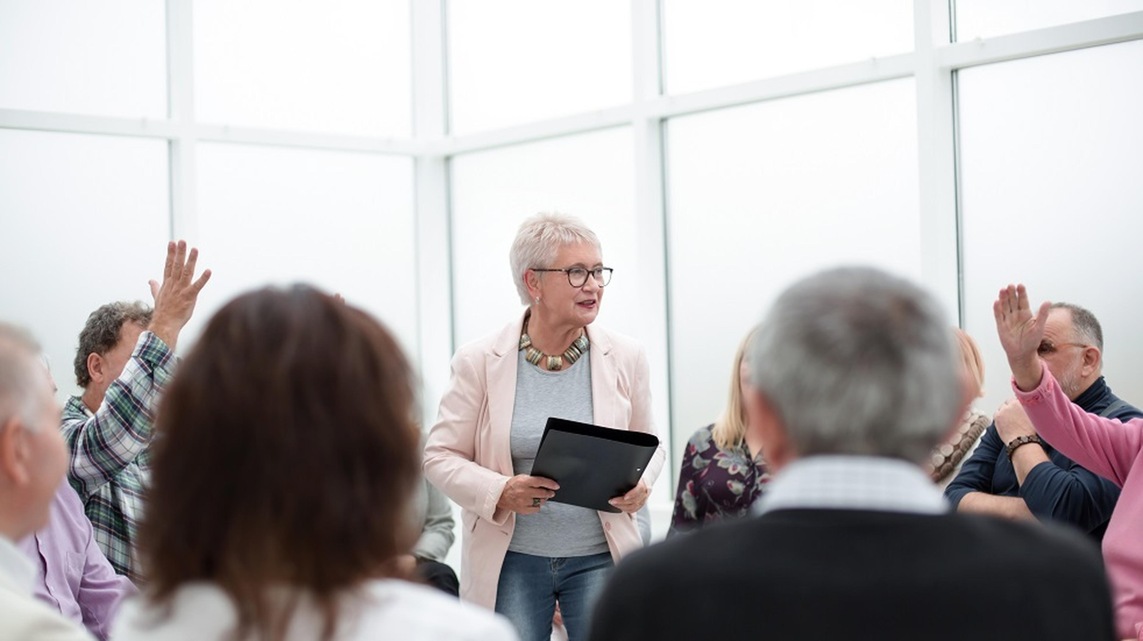 An older woman with glasses speaks to a group in a bright room