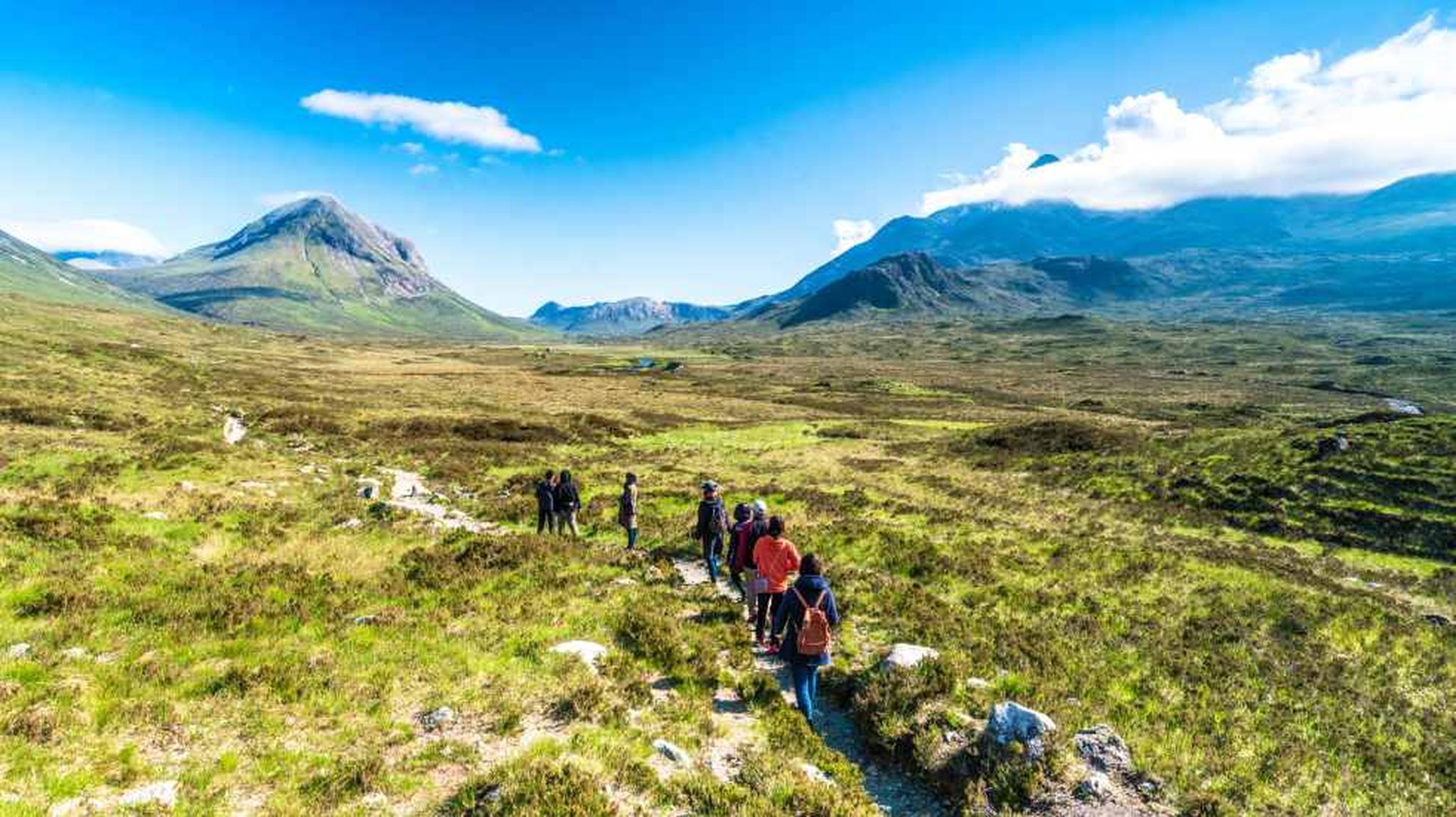 A group of hikers walking on a trail through a vast, green landscape with mountains under a clear blue sky.