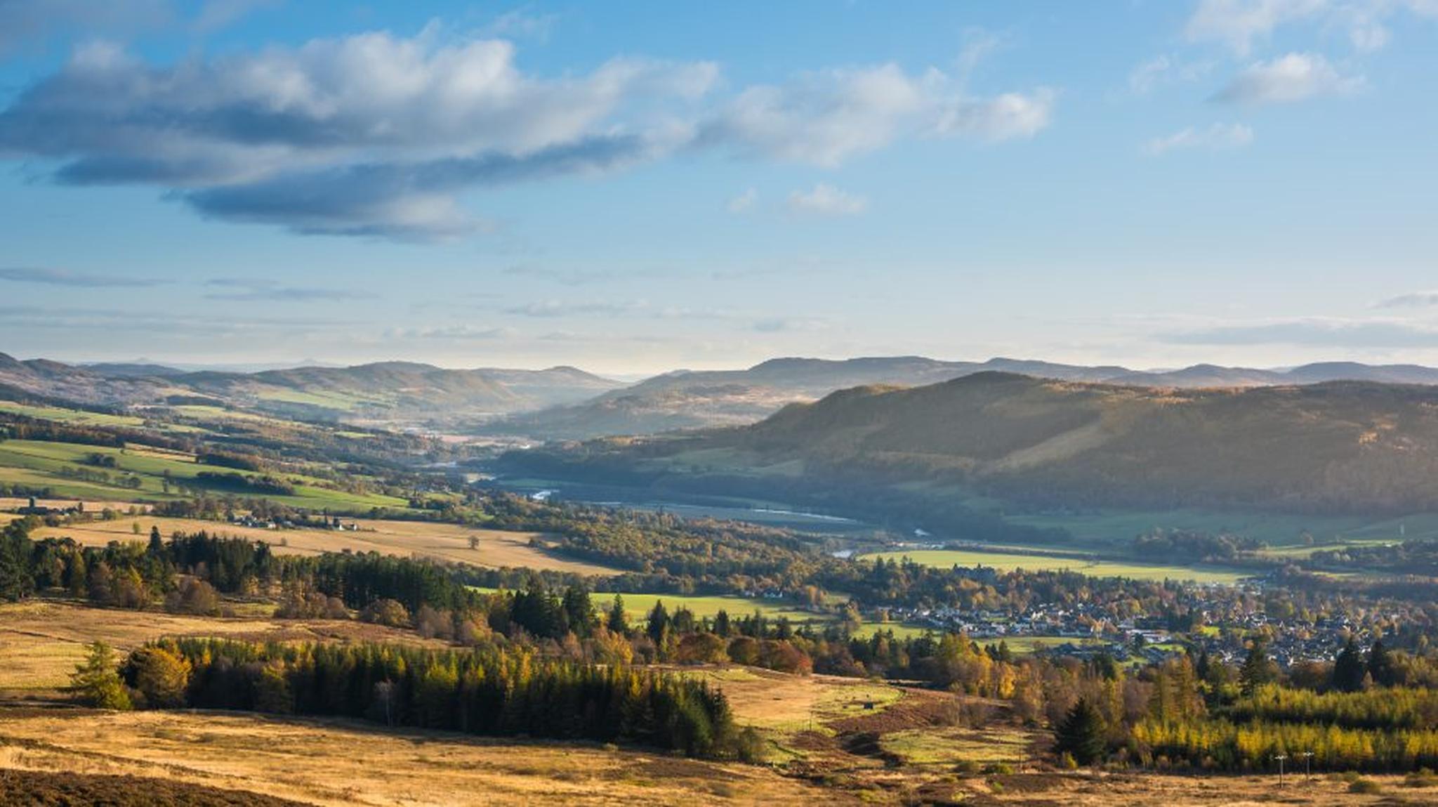 Scenic view of a valley with lush forests, rolling hills, and a river under a blue sky with scattered clouds.