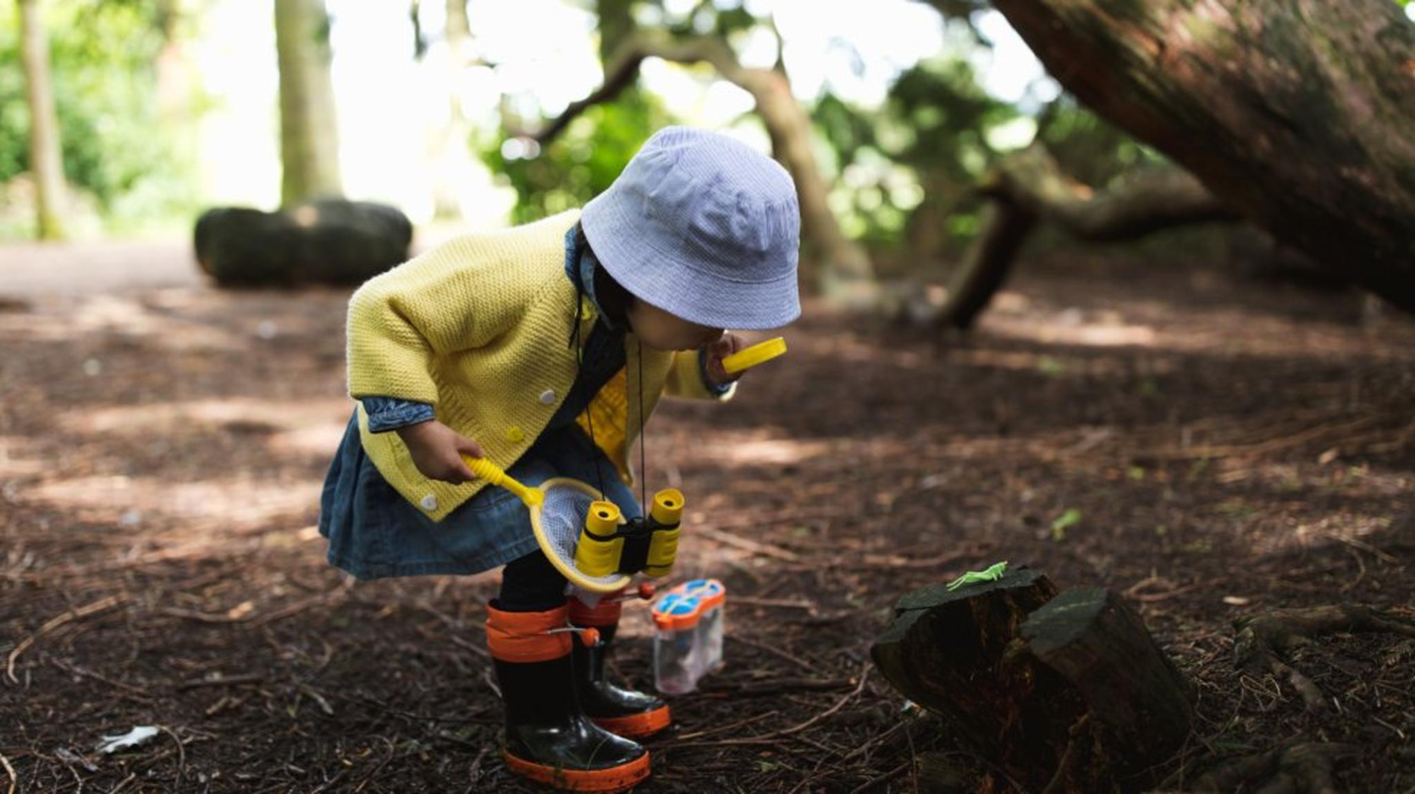 A toddler in a yellow jacket and blue hat examines a log with a magnifying glass