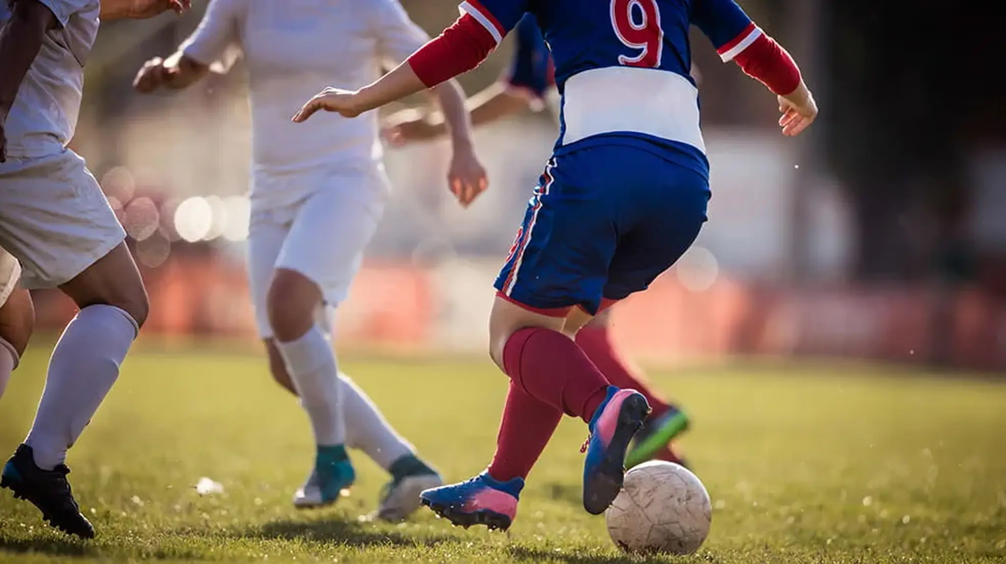 Football players in action on a grass field, with a player in blue dribbling the ball against opponents in white