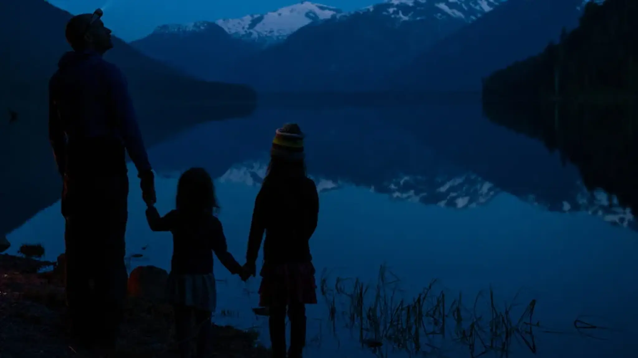 A silhouetted adult and two children hold hands by a calm lake at dusk