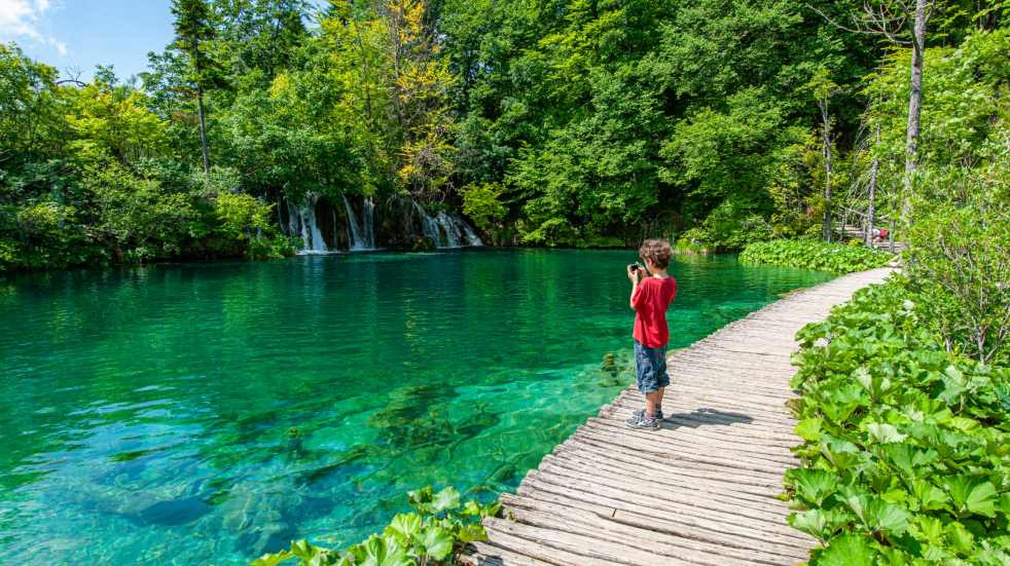 A child in a red shirt stands on a wooden path by a turquoise lake