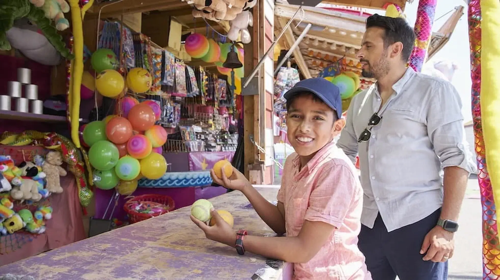 A man standing next to a boy holding balls and smiling at a carnival game booth