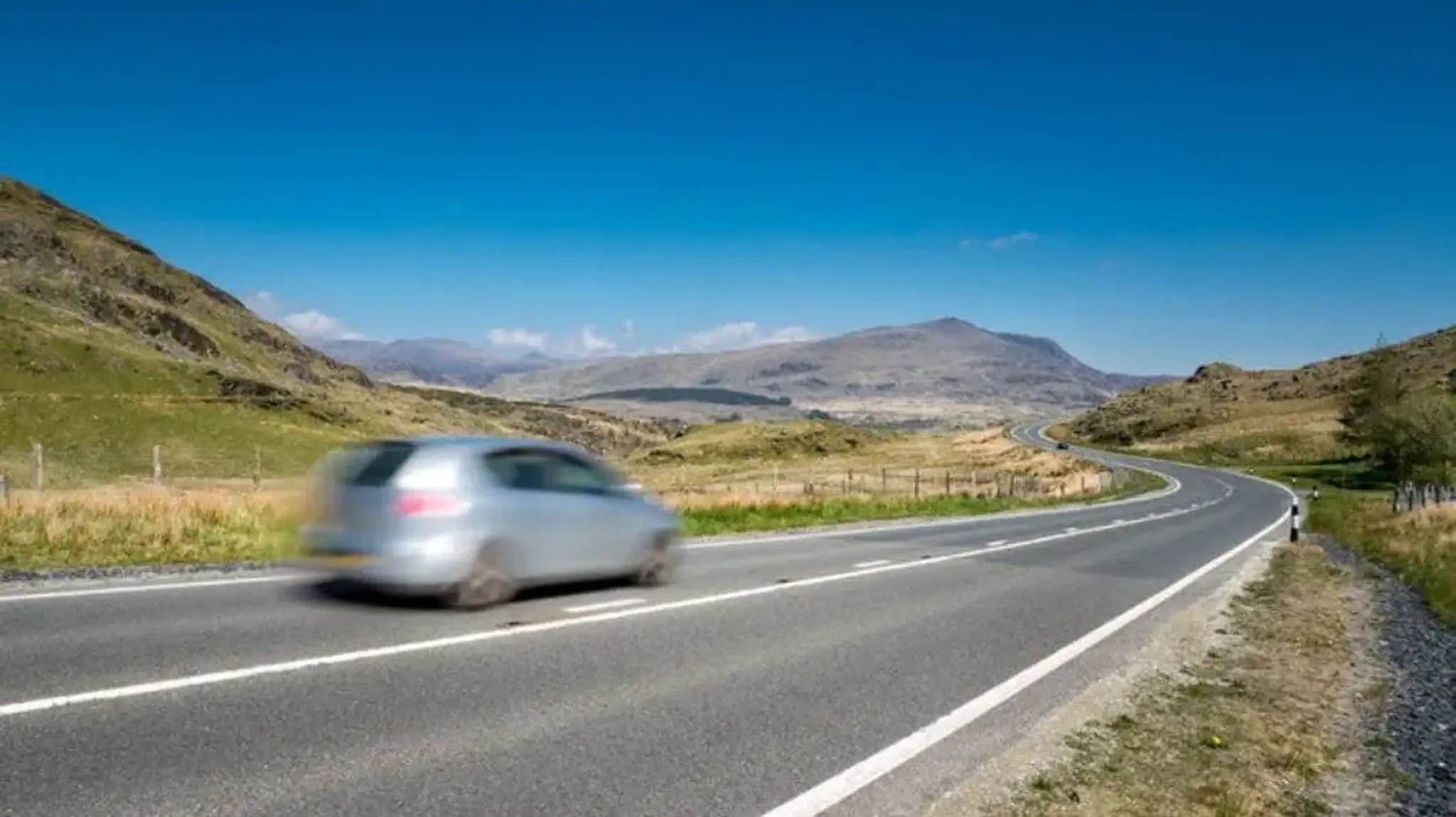 A silver car speeds along a winding rural road