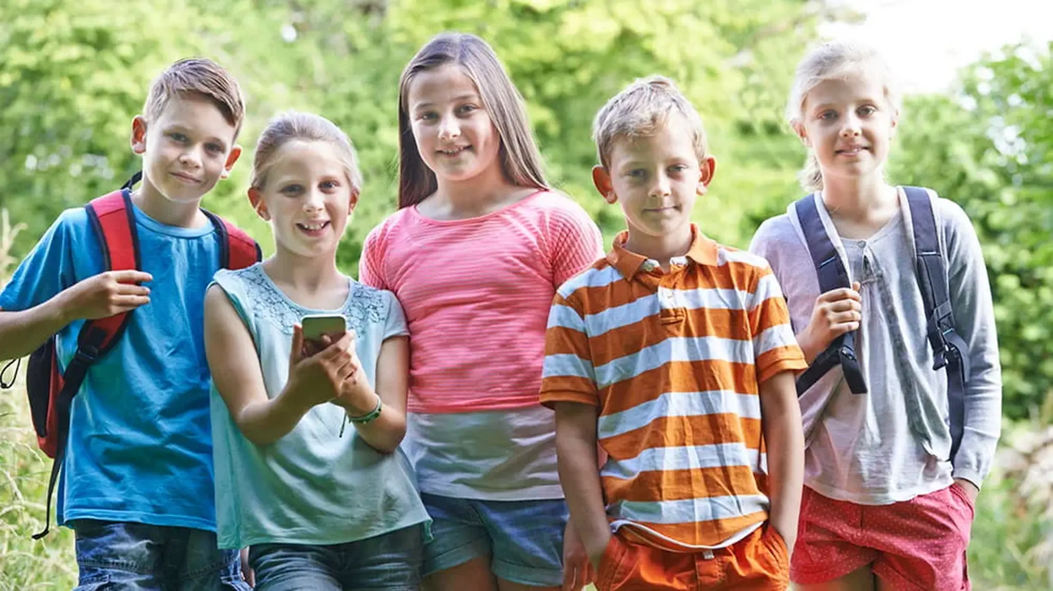 Five children smiling outdoors, standing with backpacks