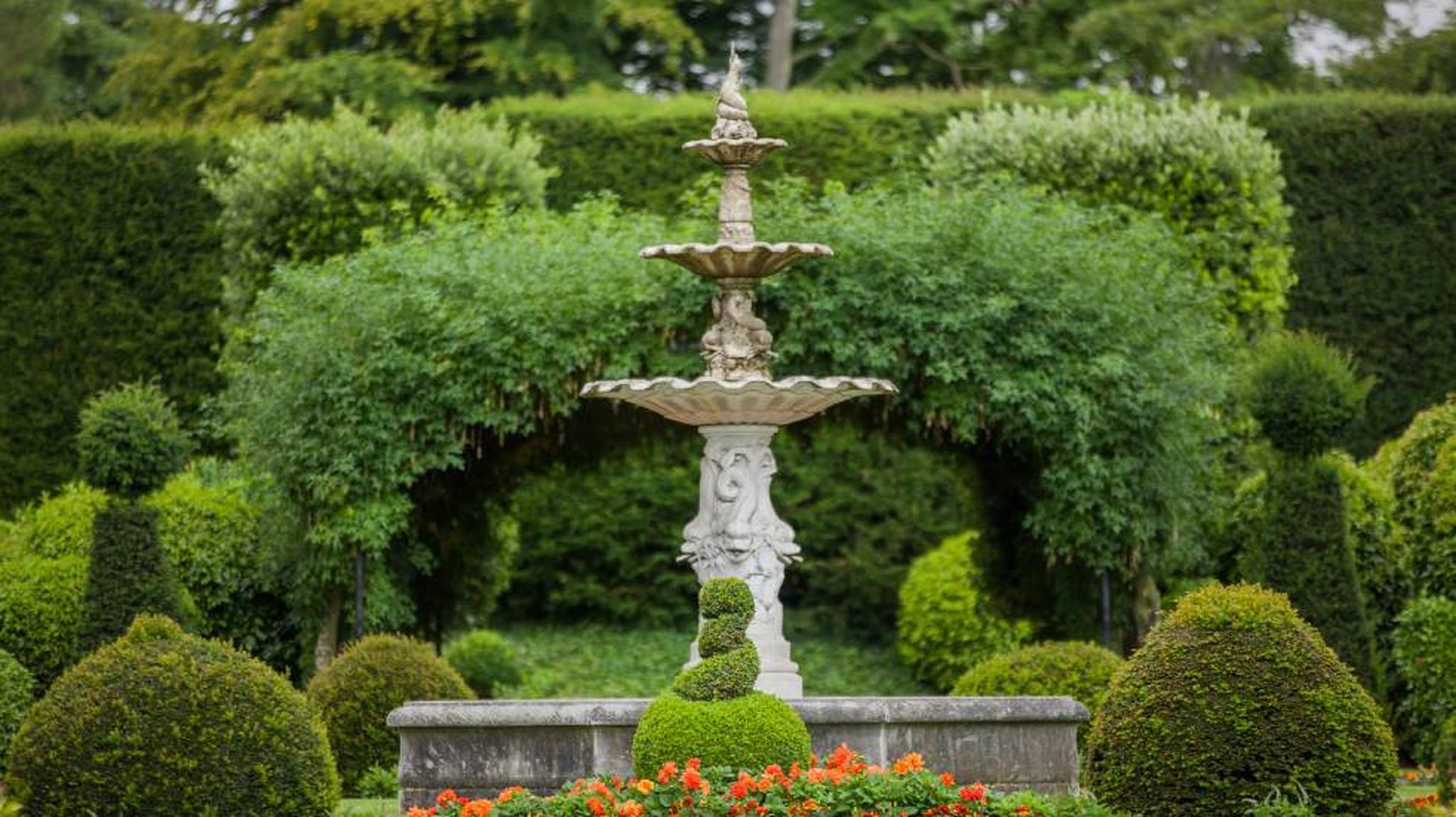 A stone fountain stands amidst greenery, surrounded by trimmed bushes