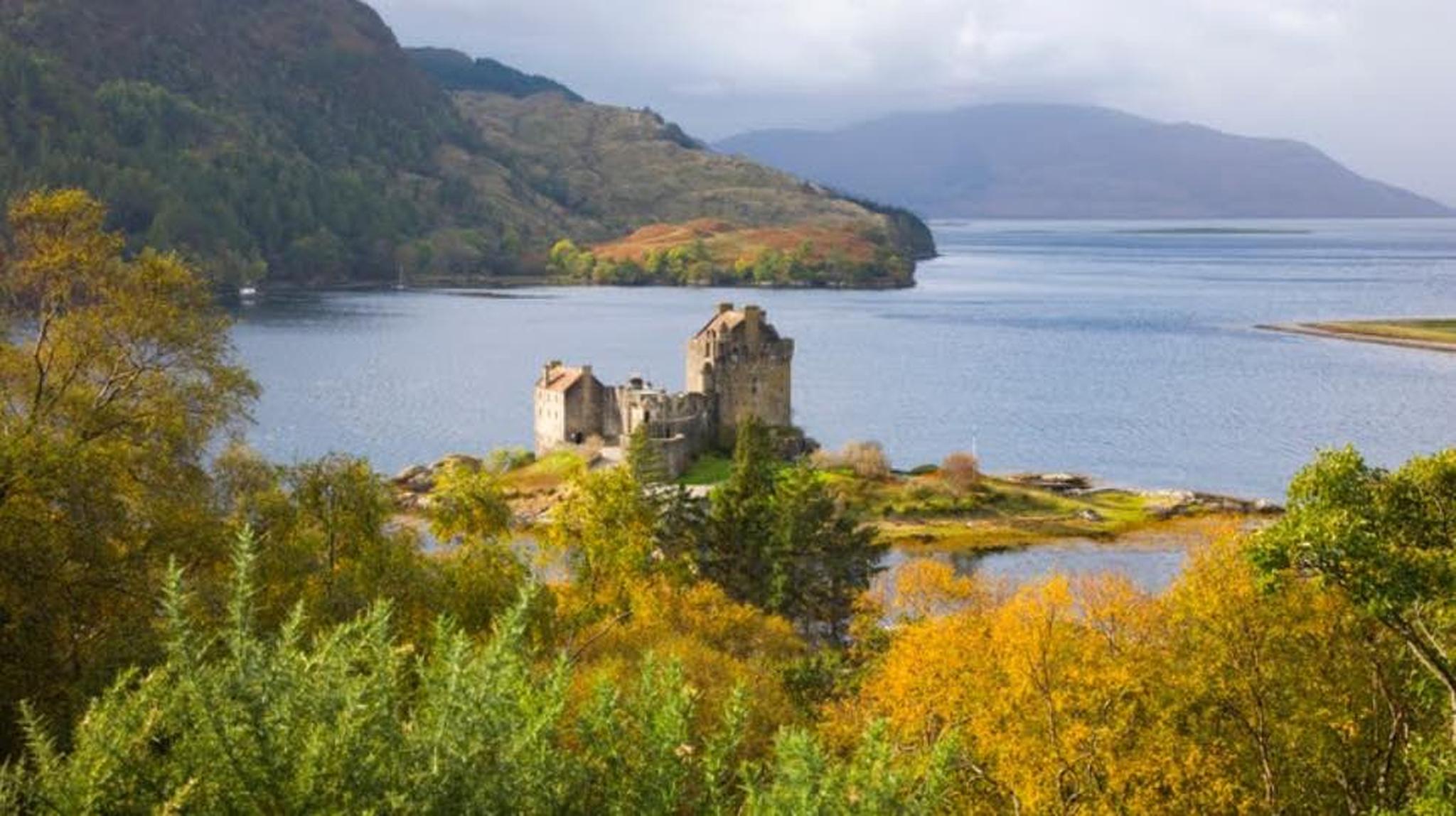 A historic stone castle sits on a small island surrounded by a serene loch, with lush green and autumnal trees in the foreground.