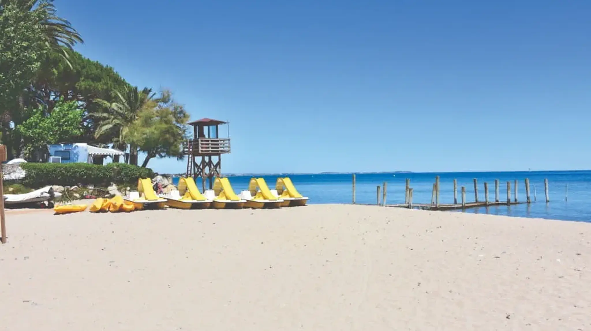 Sunny beach with yellow pedal boats lined up on the sand, a wooden pier and lifeguard tower