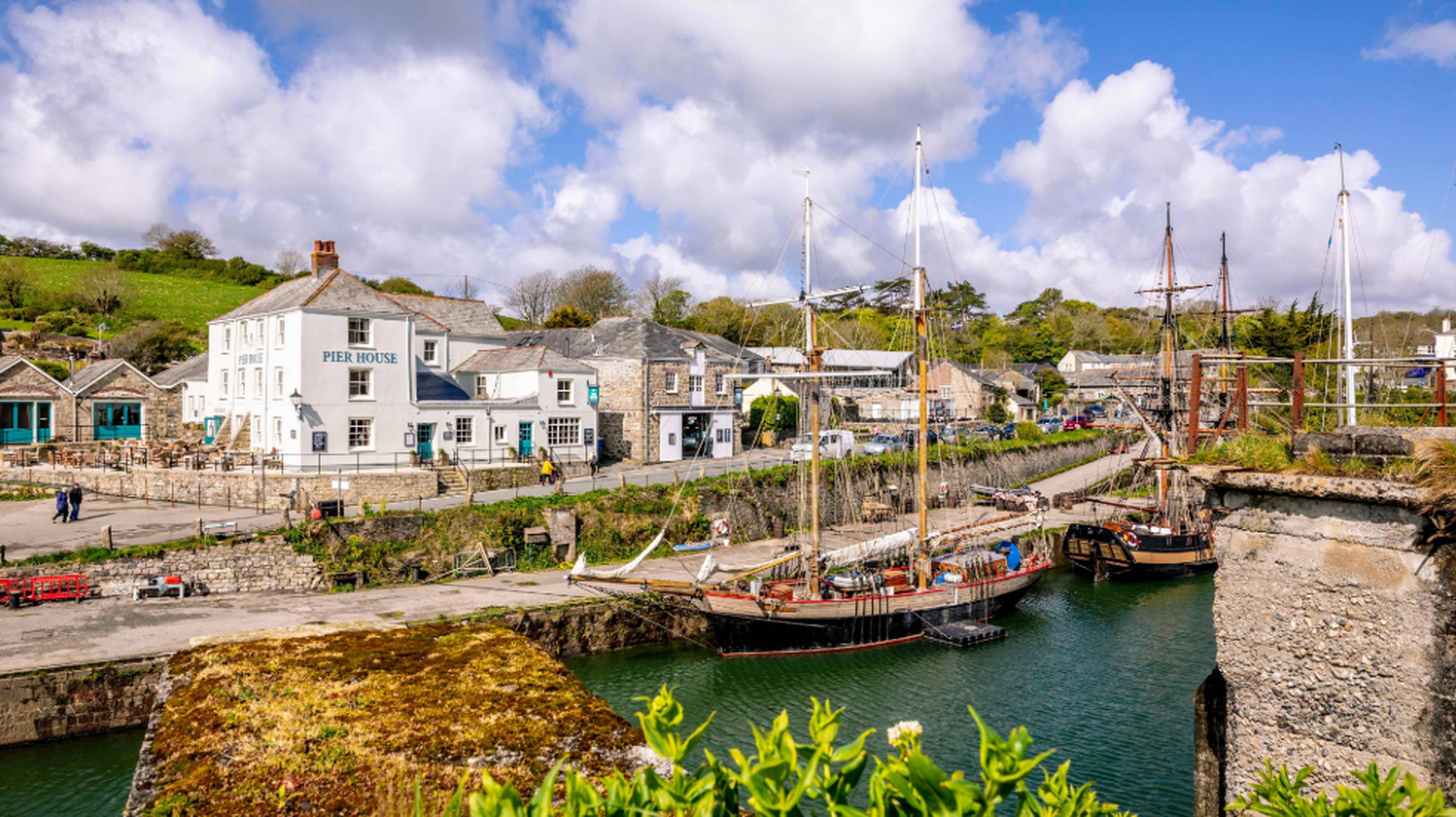A picturesque harbor with sailboats, stone buildings, lush greenery, and a cloudy blue sky in a quaint seaside village.