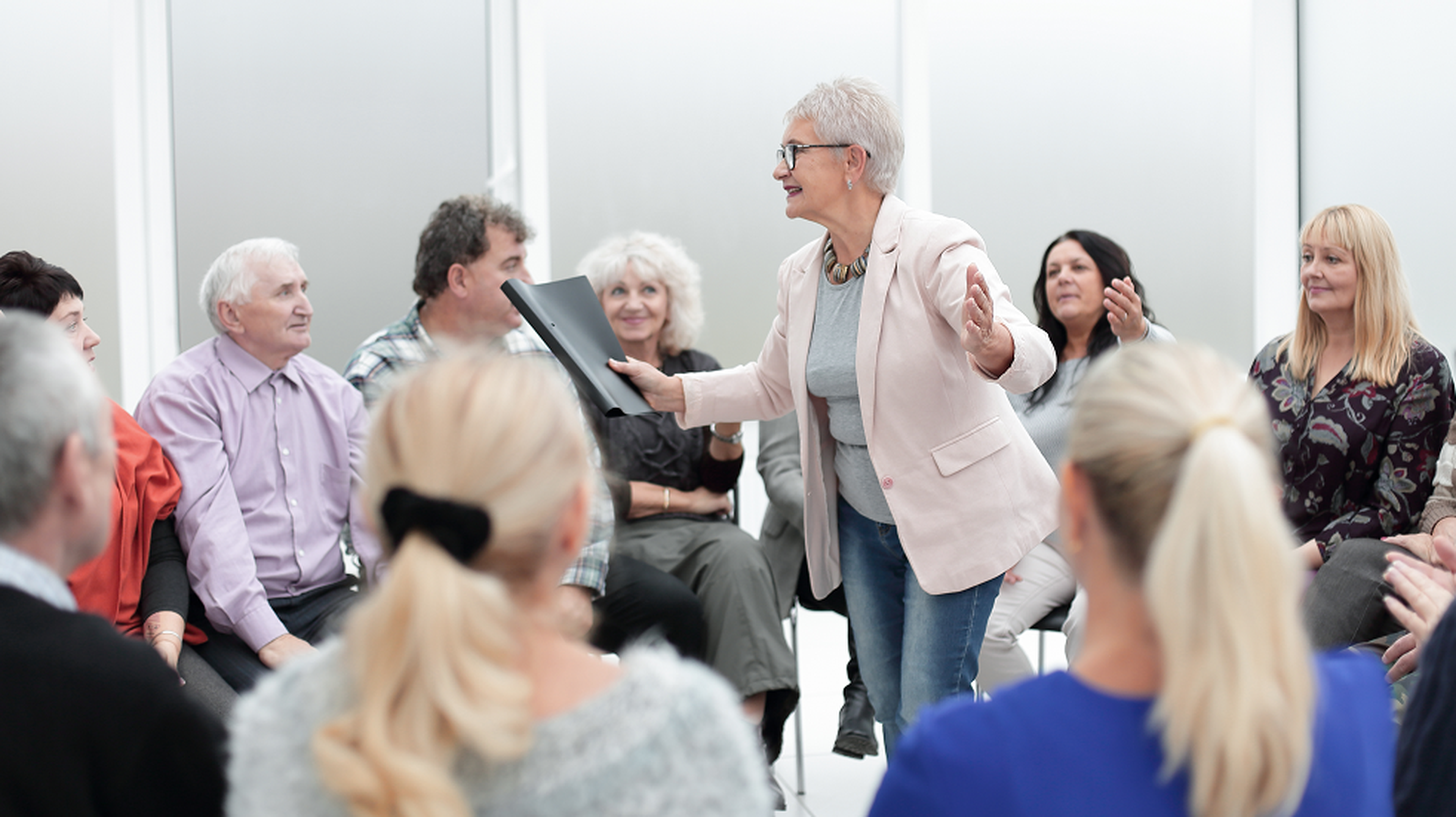 A woman in a pink blazer leads a lively discussion with an engaged group of people seated in a circle.