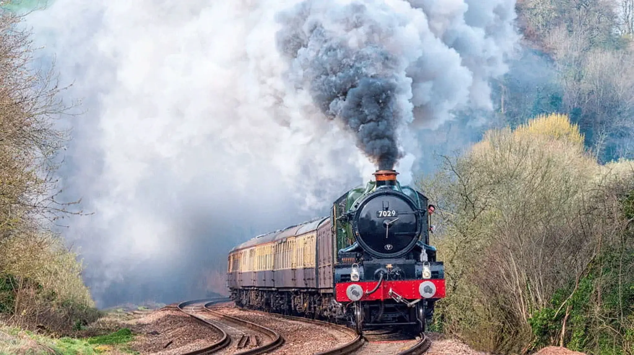 Steam train with billowing smoke travels along a curved track surrounded by greenery