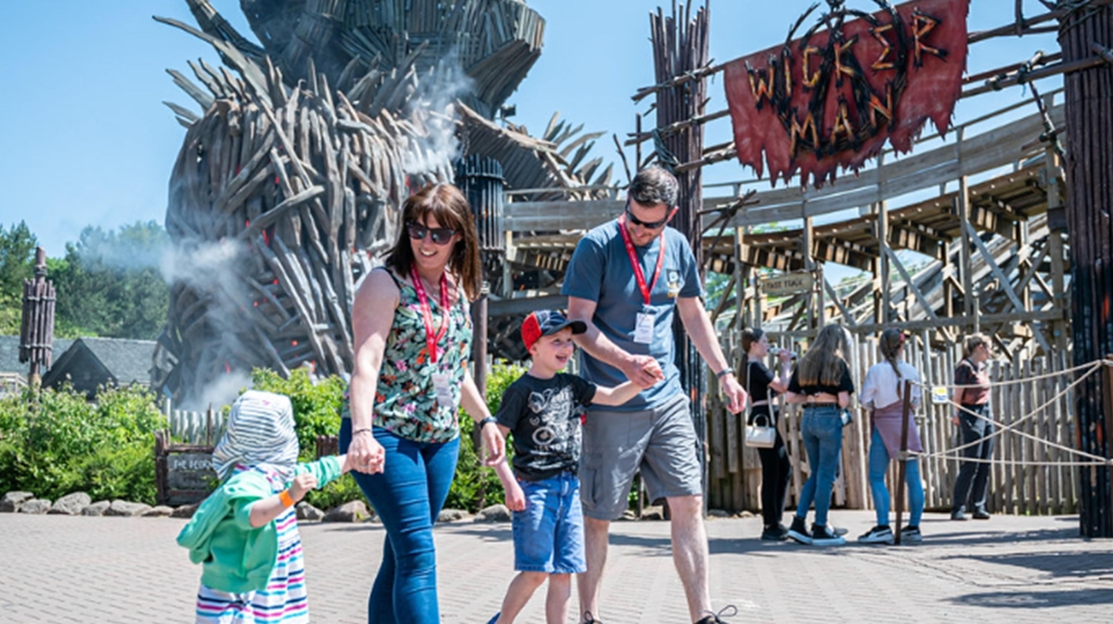 Family walking together at an amusement park, near a large wooden structure and "Wicker Man" sign, on a sunny day.