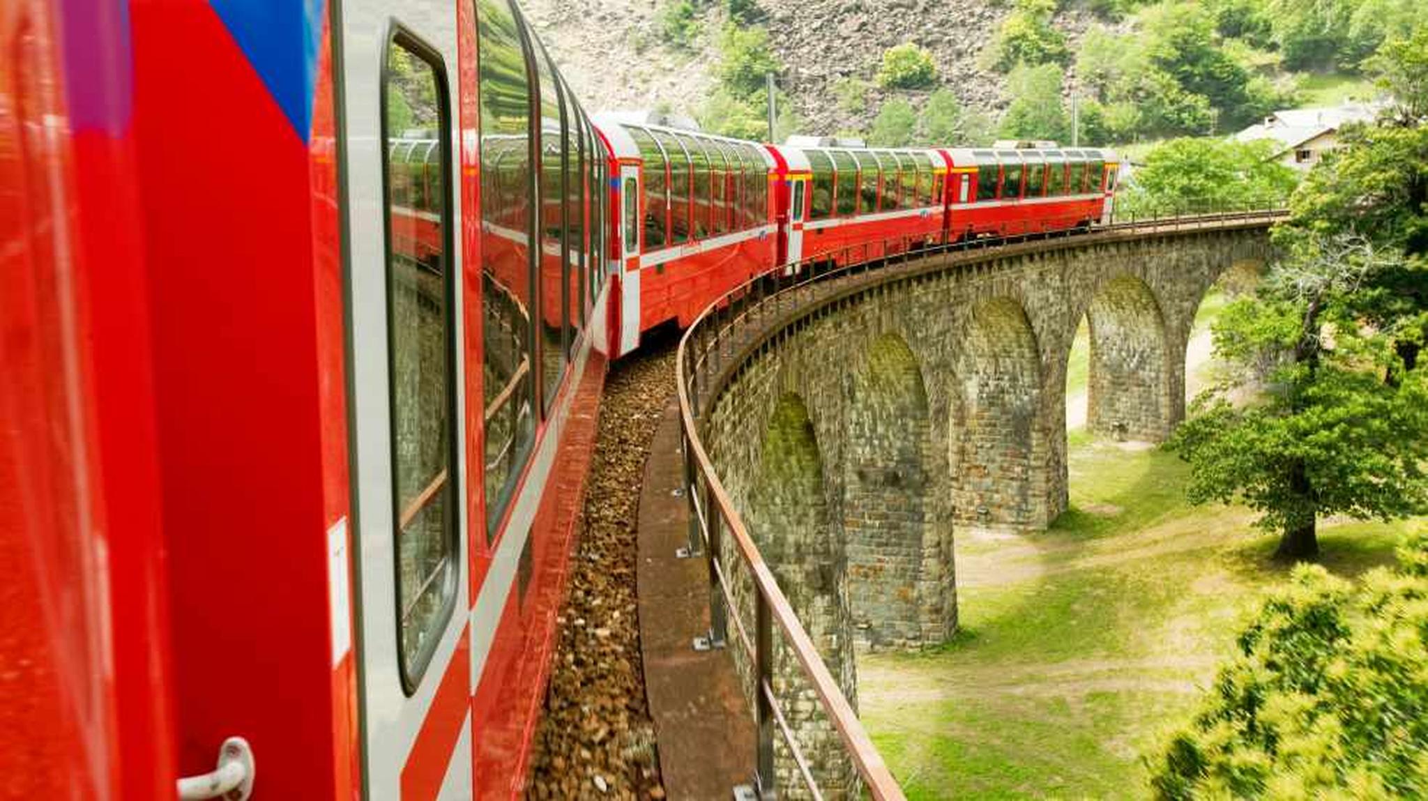 Red train on a curved stone viaduct traverses a green landscape