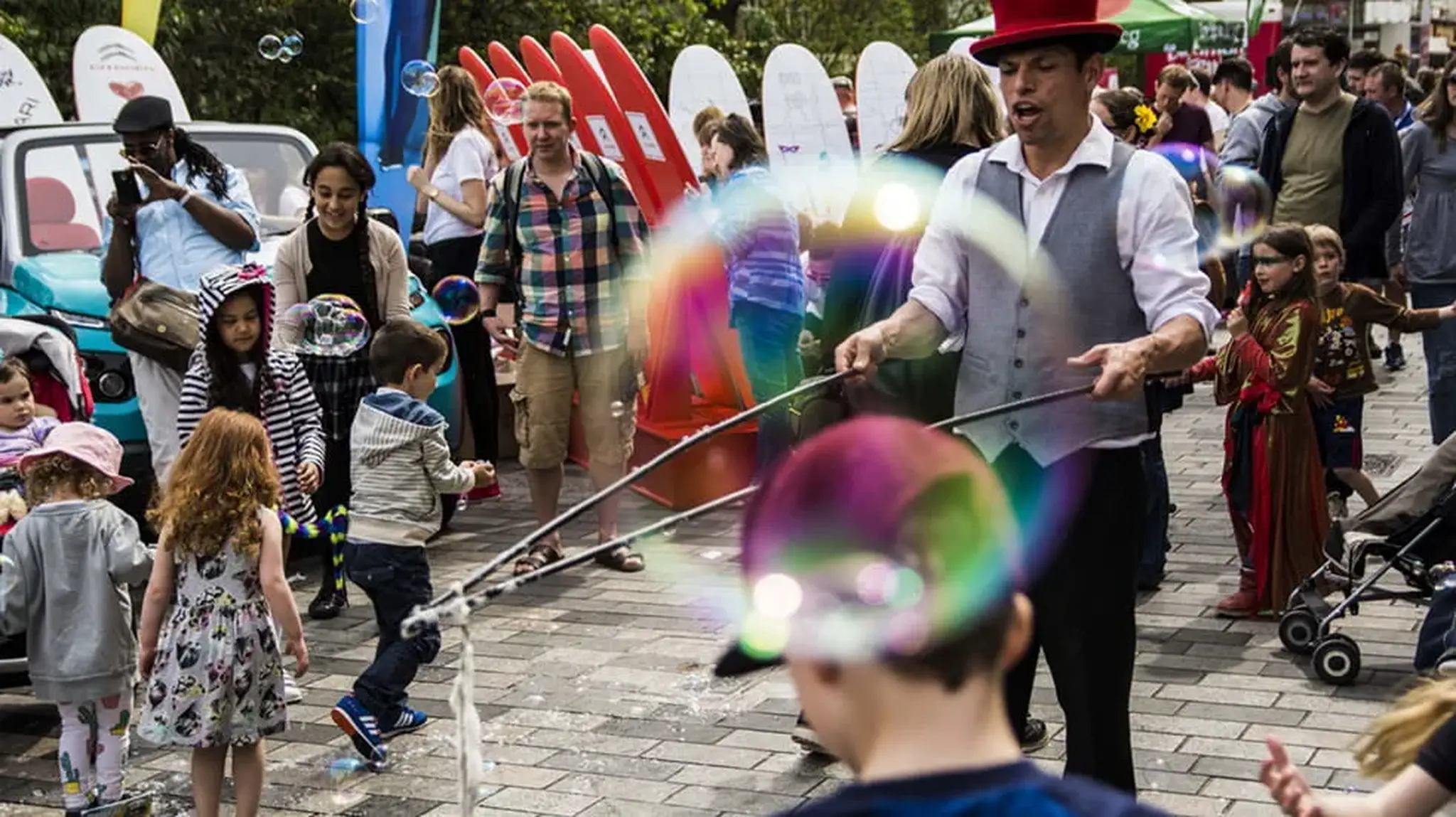 A street performer in a red hat and vest creates large bubbles, surrounded by a crowd