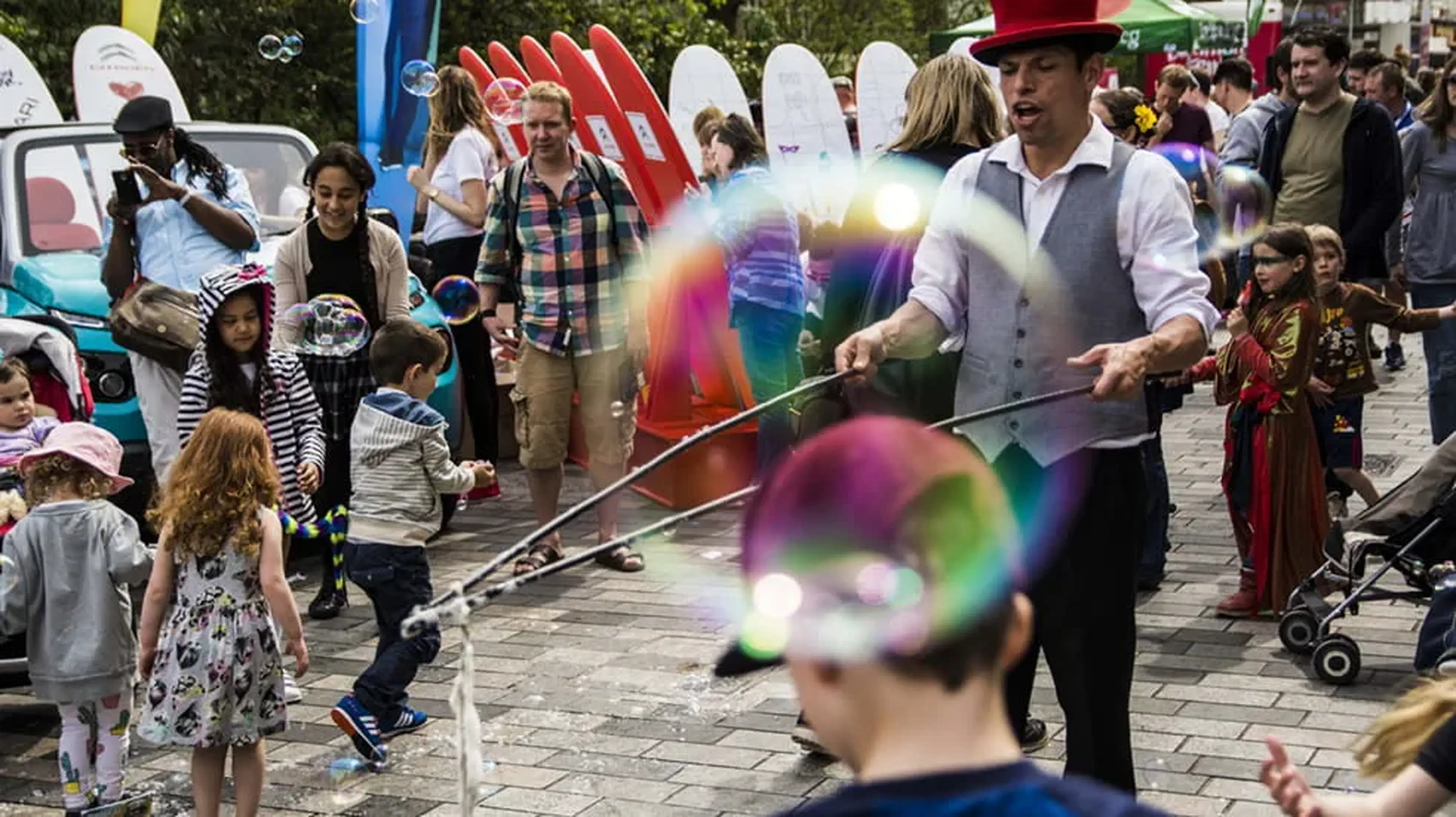 A street performer in a red hat and vest creates large bubbles, surrounded by a crowd