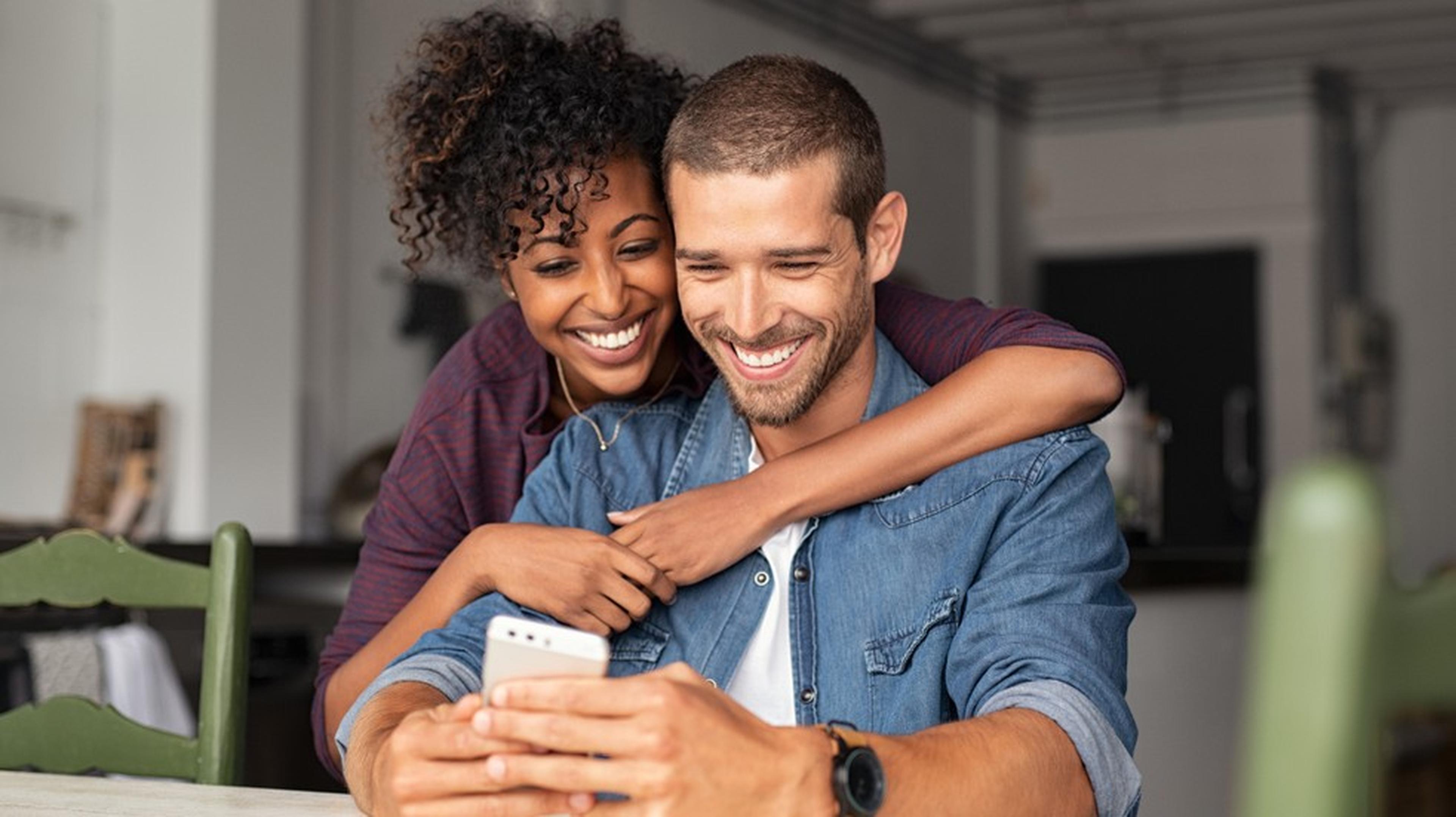 A smiling couple sits closely at a table, looking at a smartphone together