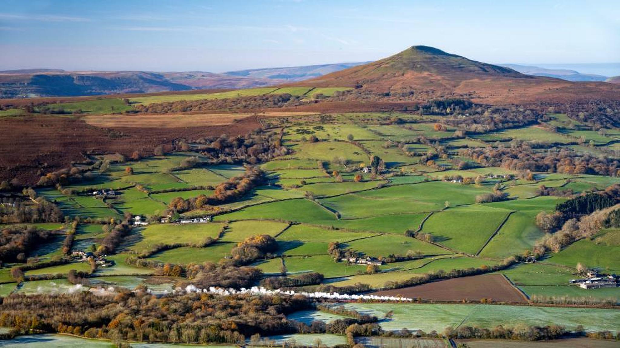 Aerial view of a lush, green valley with patchwork fields, a winding river, and a prominent hill in the background under a clear blue sky.