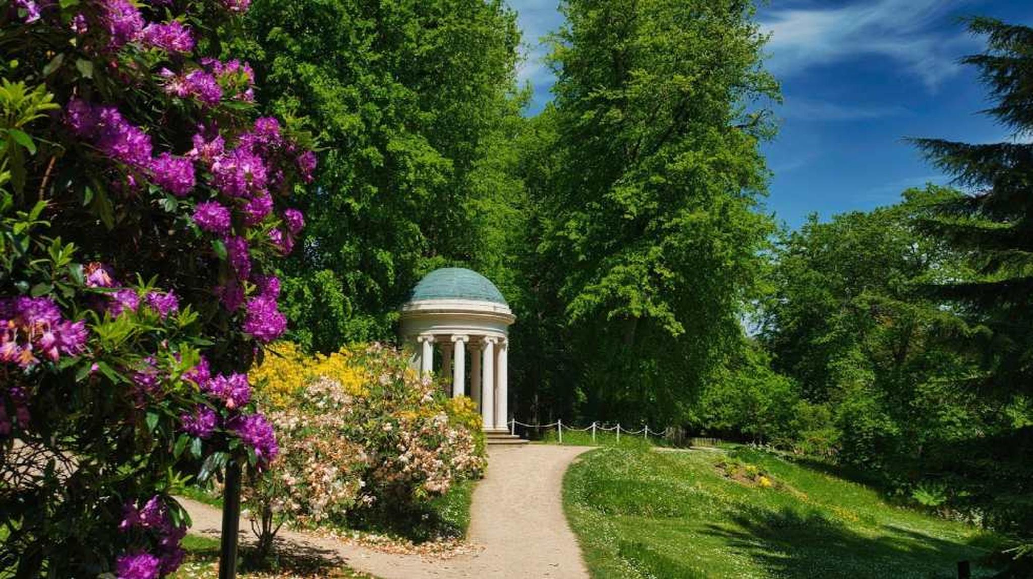 A small, round gazebo with columns sits among greenery and blooming flowers