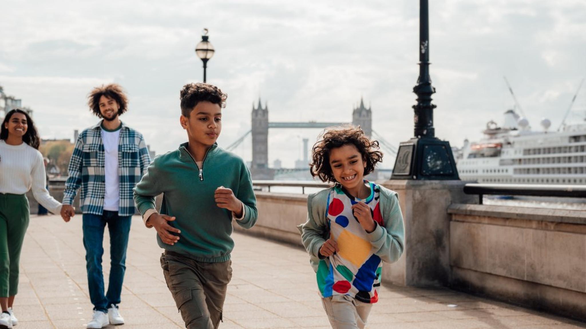 Two children smile as they run along a riverside walkway, with a couple walking behind them