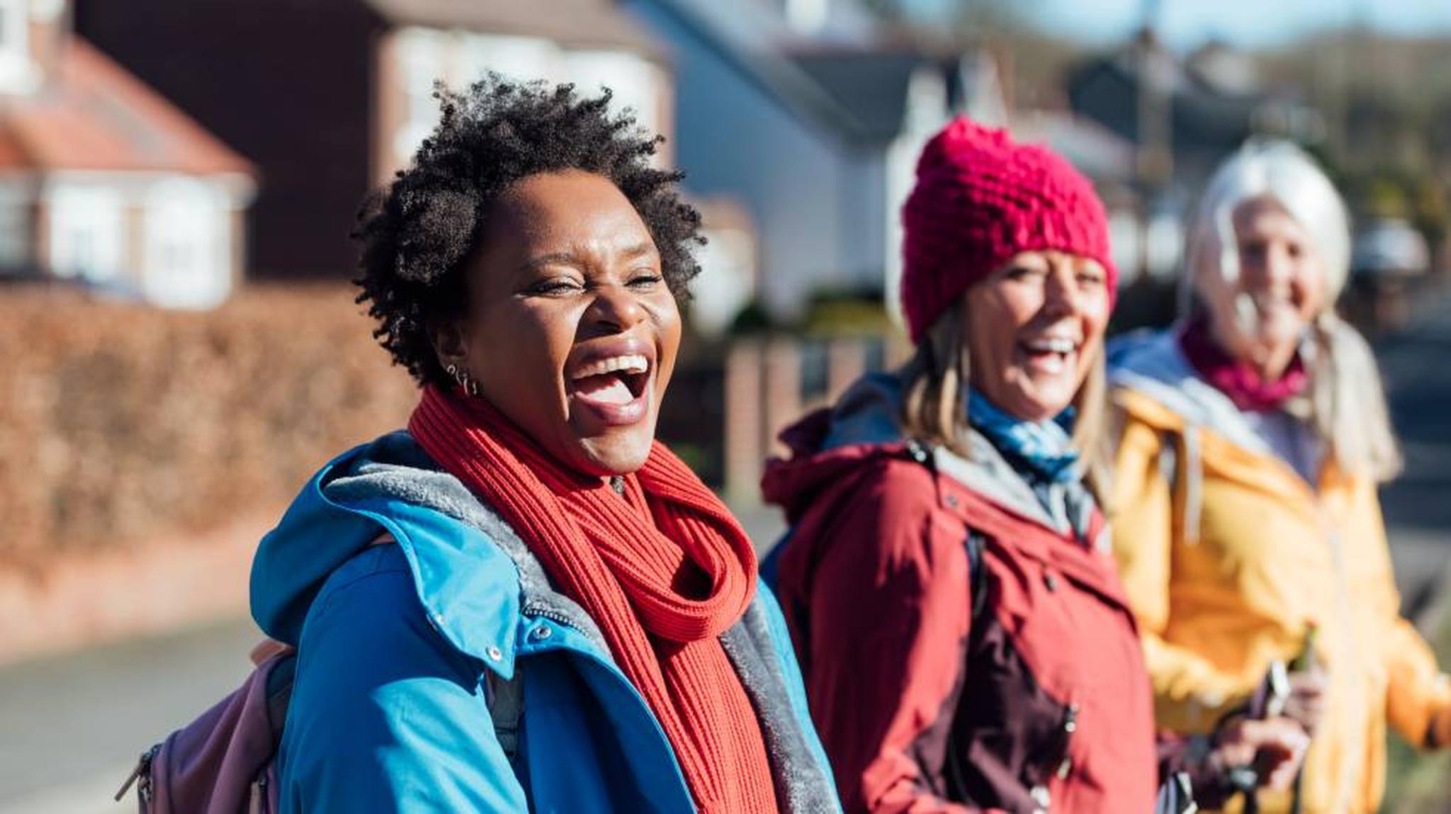 Three women in colorful winter clothing laugh joyfully while walking outdoors in a suburban neighborhood.