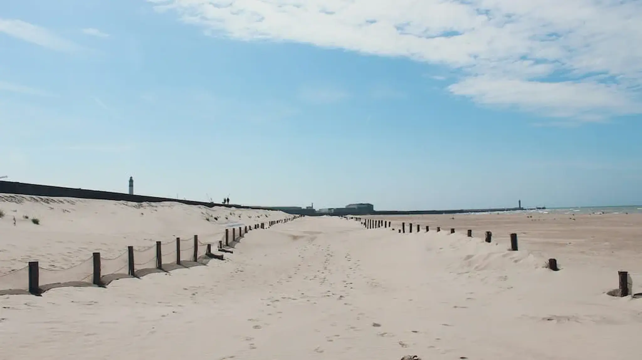Sandy beach path with wooden posts and two distant lighthouses visible on the horizon