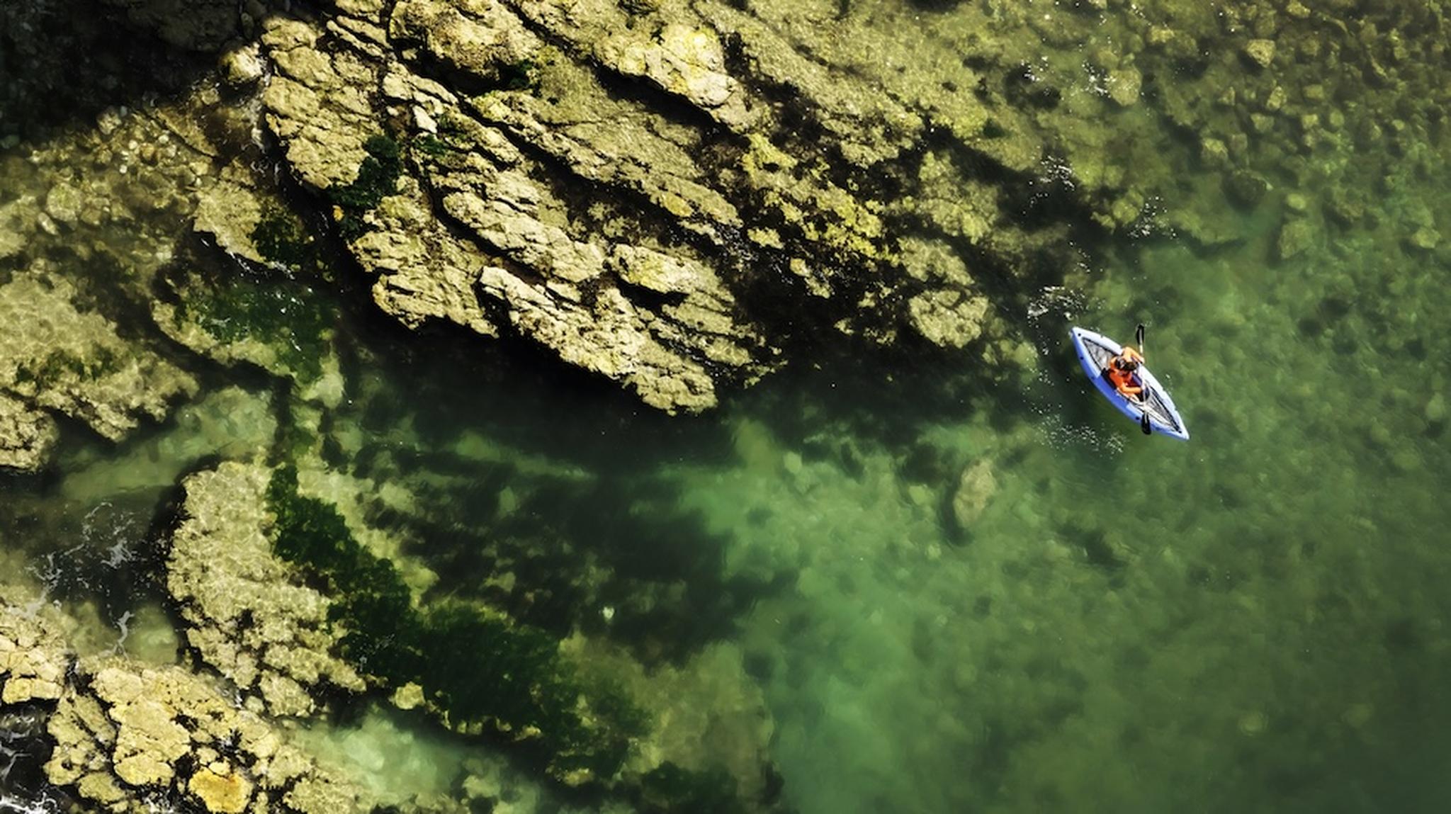 Aerial view of a person in a kayak paddling near rocky cliffs in clear, turquoise water.