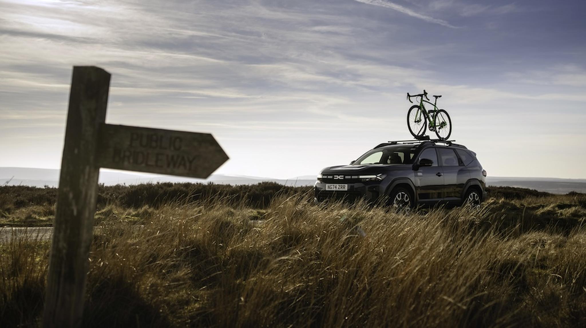 SUV with a bike on the roof parked on a grassy landscape; a wooden public footpath sign in the foreground points left.