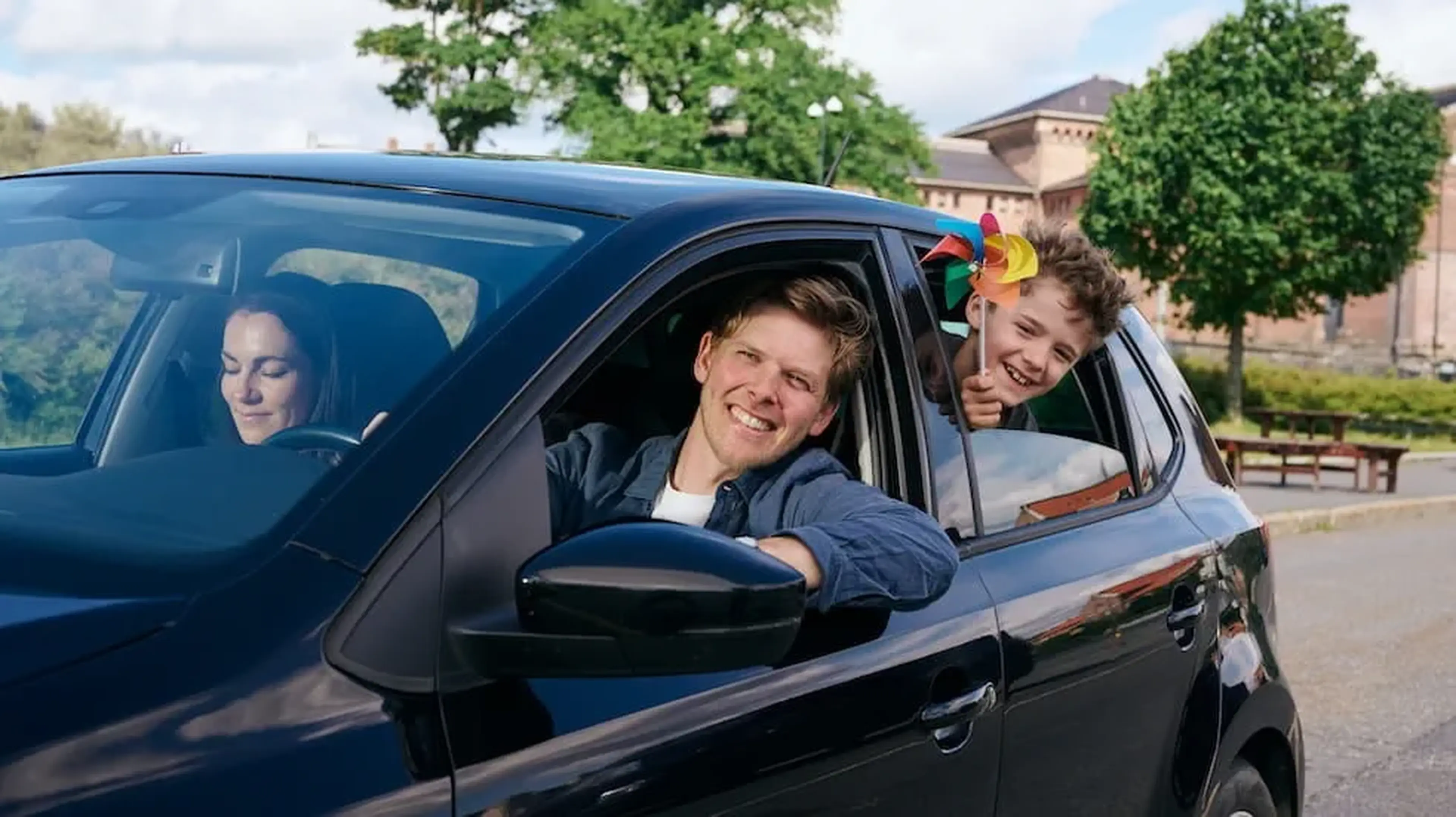 A car with a woman driving, a smiling man leaning out of the window and a boy holding a pinwheel