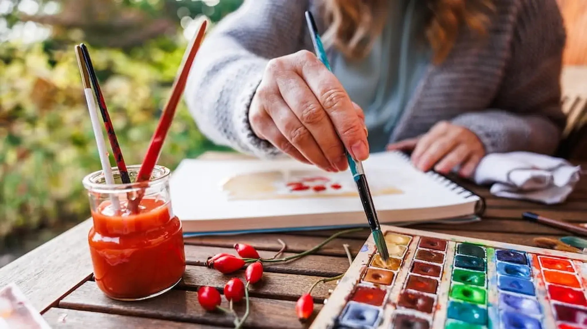 A person painting outdoors with watercolours, using a palette and jar with brushes