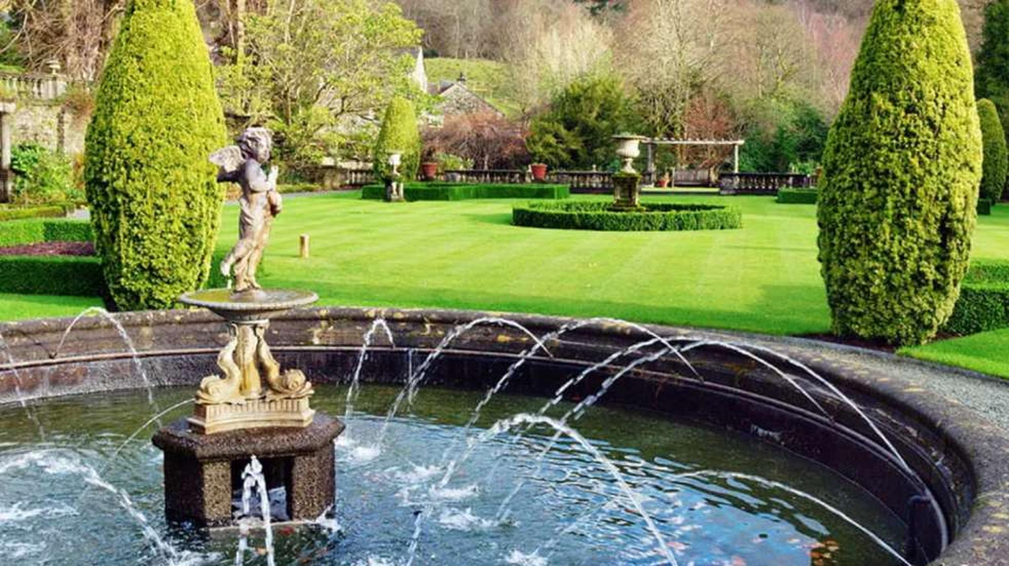 A landscaped garden with a central fountain, manicured lawns, tall trimmed trees, and statues