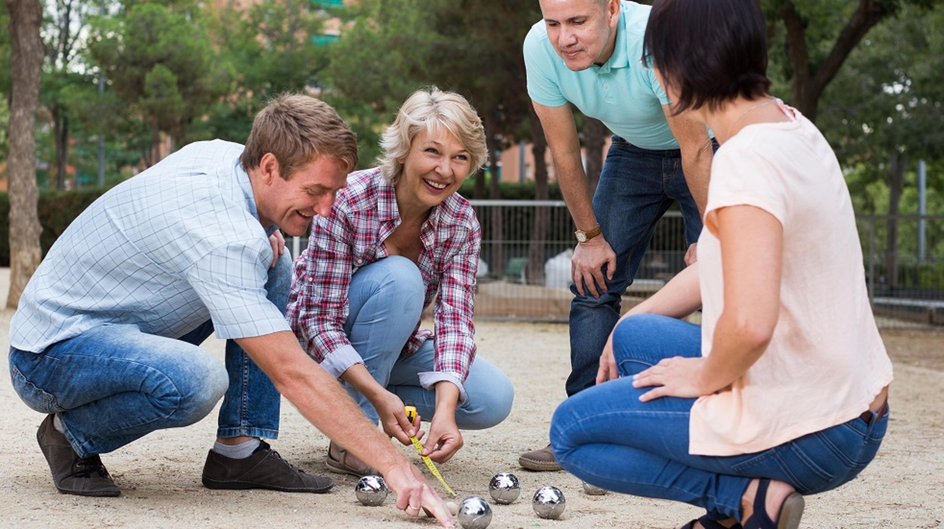 Four people are playing pétanque outdoors, measuring the distance between metal balls on a sandy surface, smiling and engaged.
