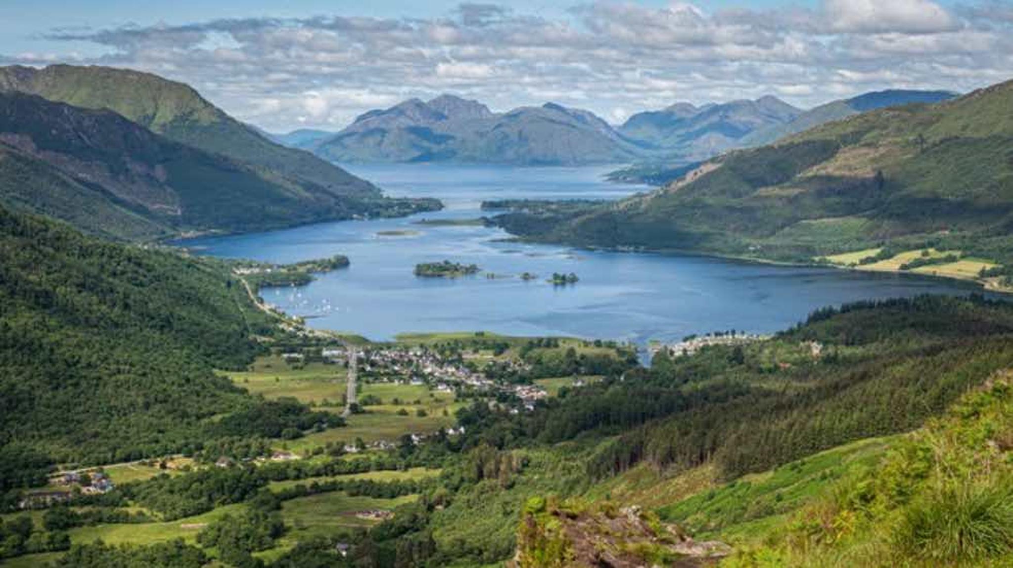 Scenic view of a lake surrounded by lush green hills and mountains, with a small village nestled along the shoreline.