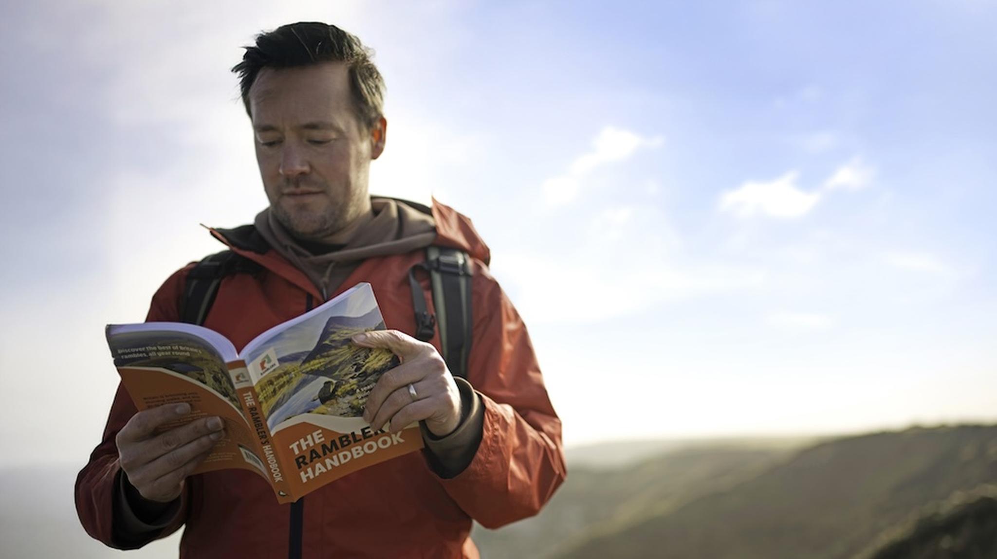 Man in a red jacket stands on a hilltop, reading "The Rambler's Handbook,"