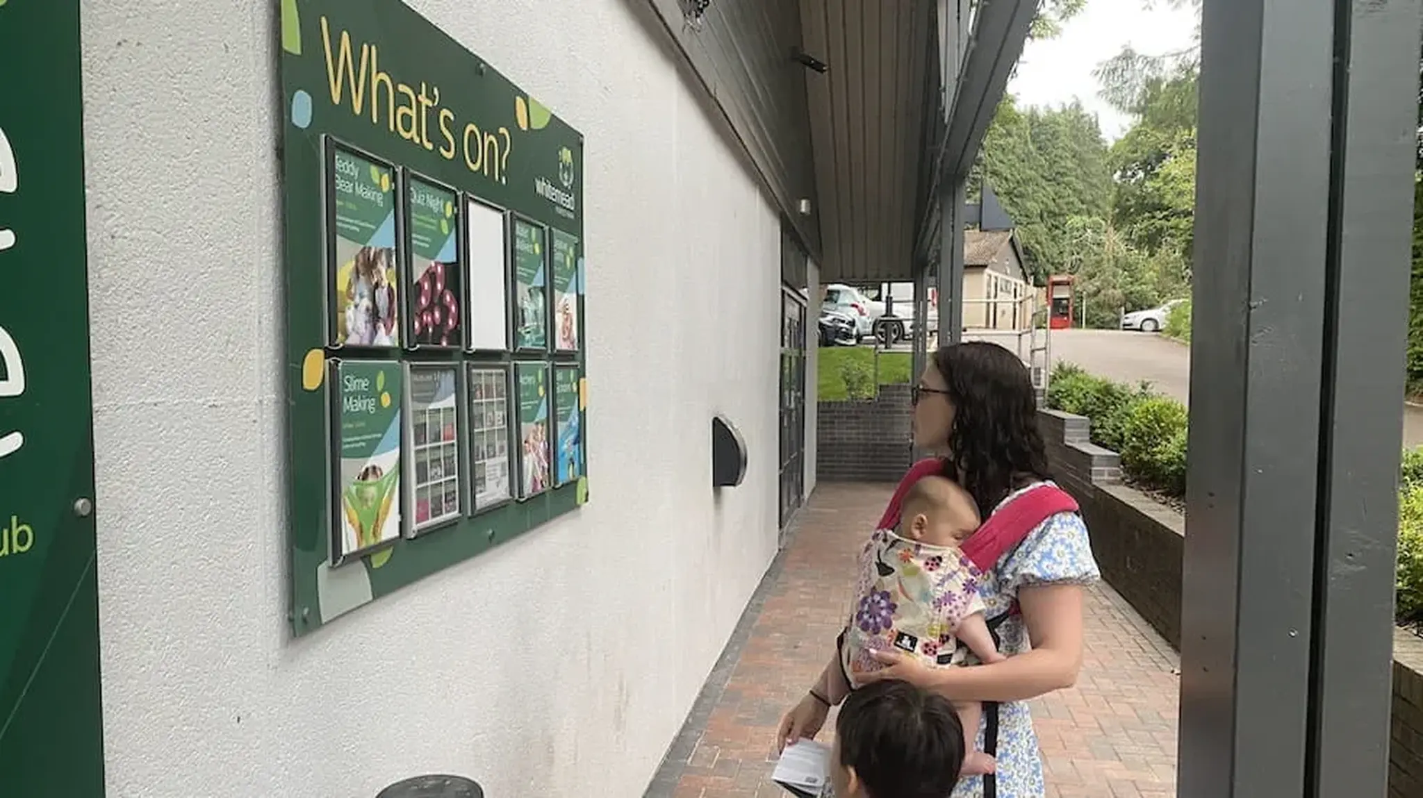 A woman with a sleeping baby in a carrier and a child holding her hand looks at a "What's on?" board outside a building