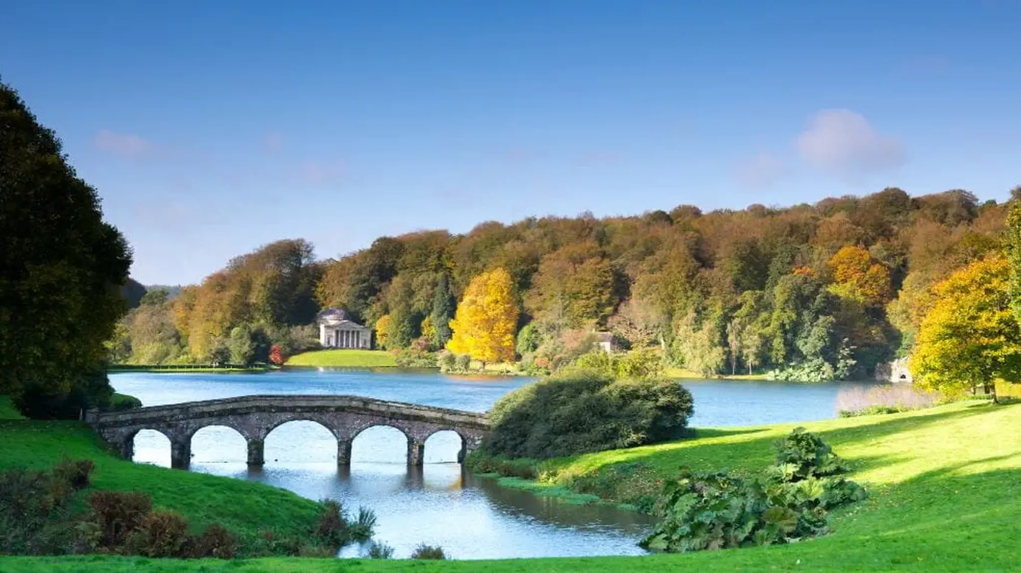 Scenic landscape of a stone bridge over a lake with autumn trees and a classical building in the background