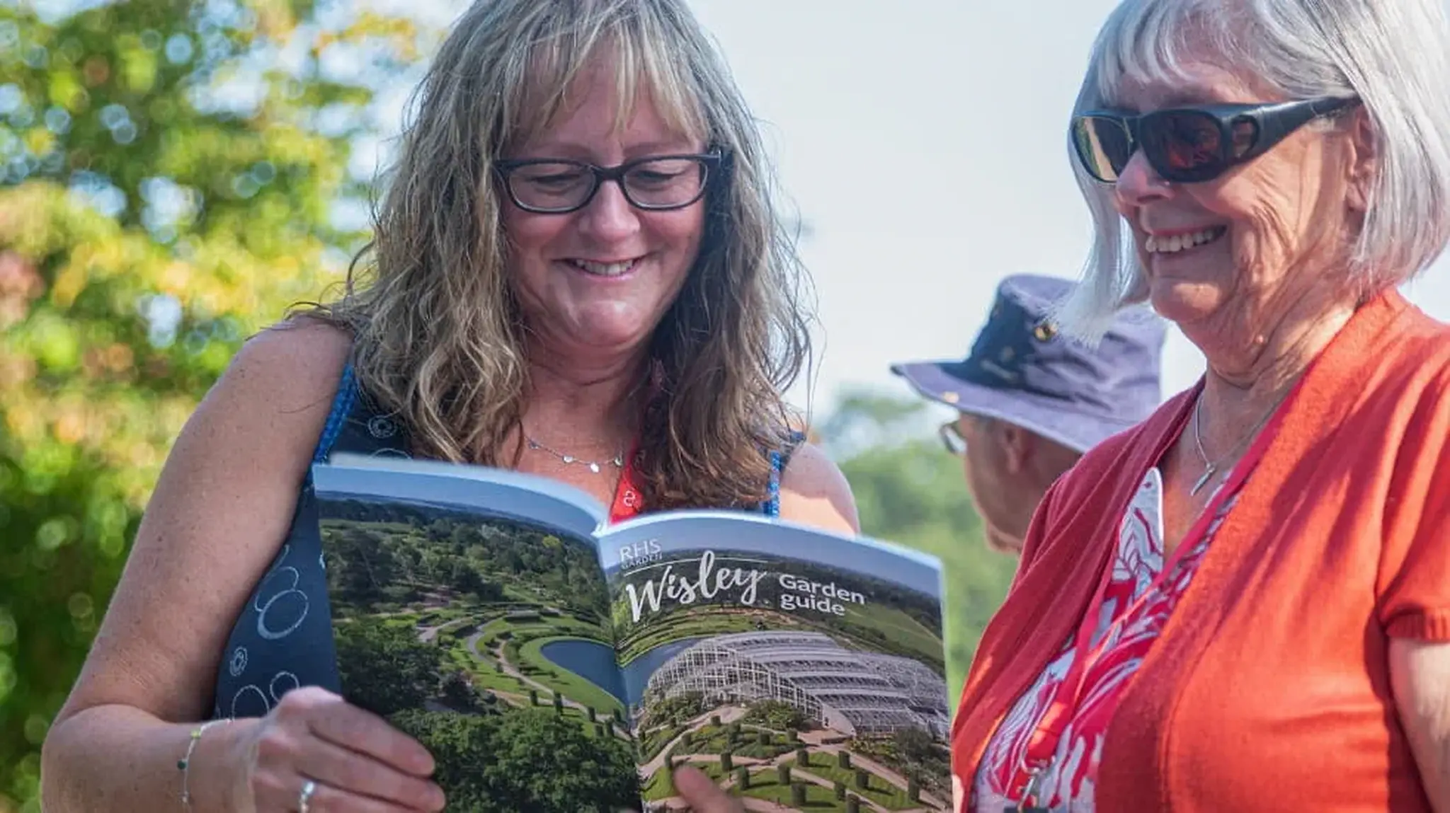 Two women smiling, reading an RHS Wisley garden guide outdoors