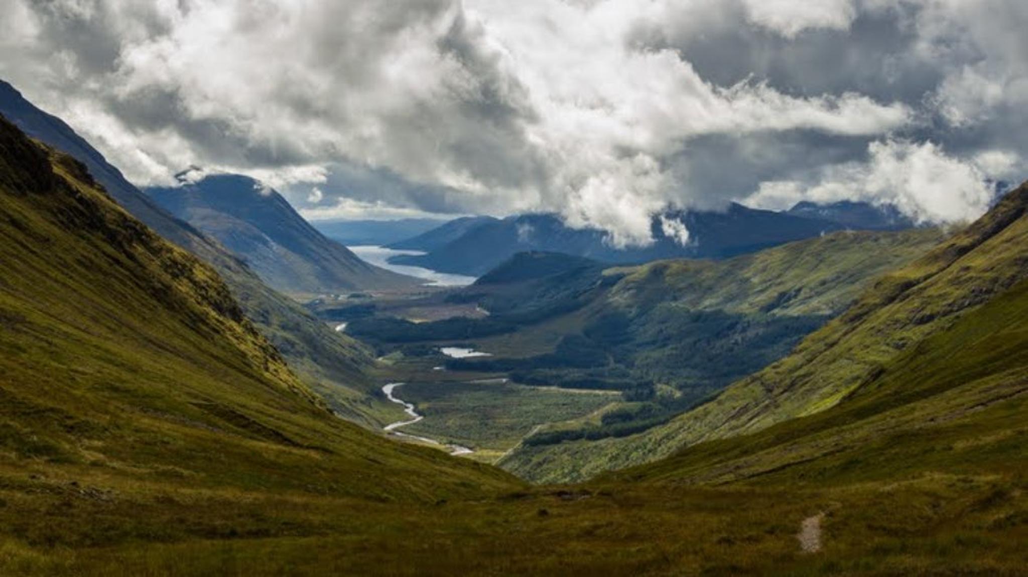 A lush, green valley with a winding river under dramatic, cloud-filled skies, flanked by mountainous terrain.