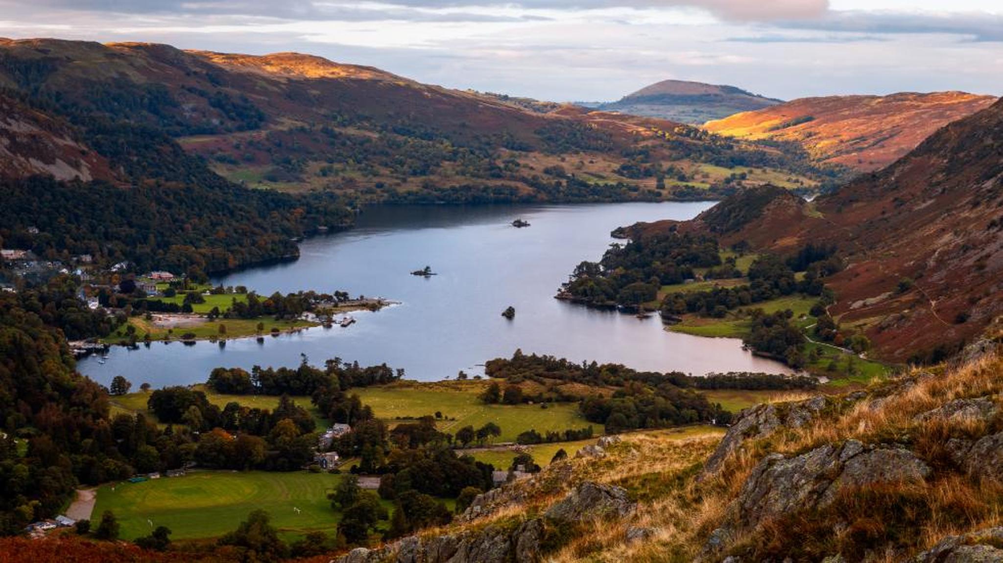 A scenic view of a lake surrounded by hills and trees, with patches of sunlight highlighting the landscape, under a partly cloudy sky.