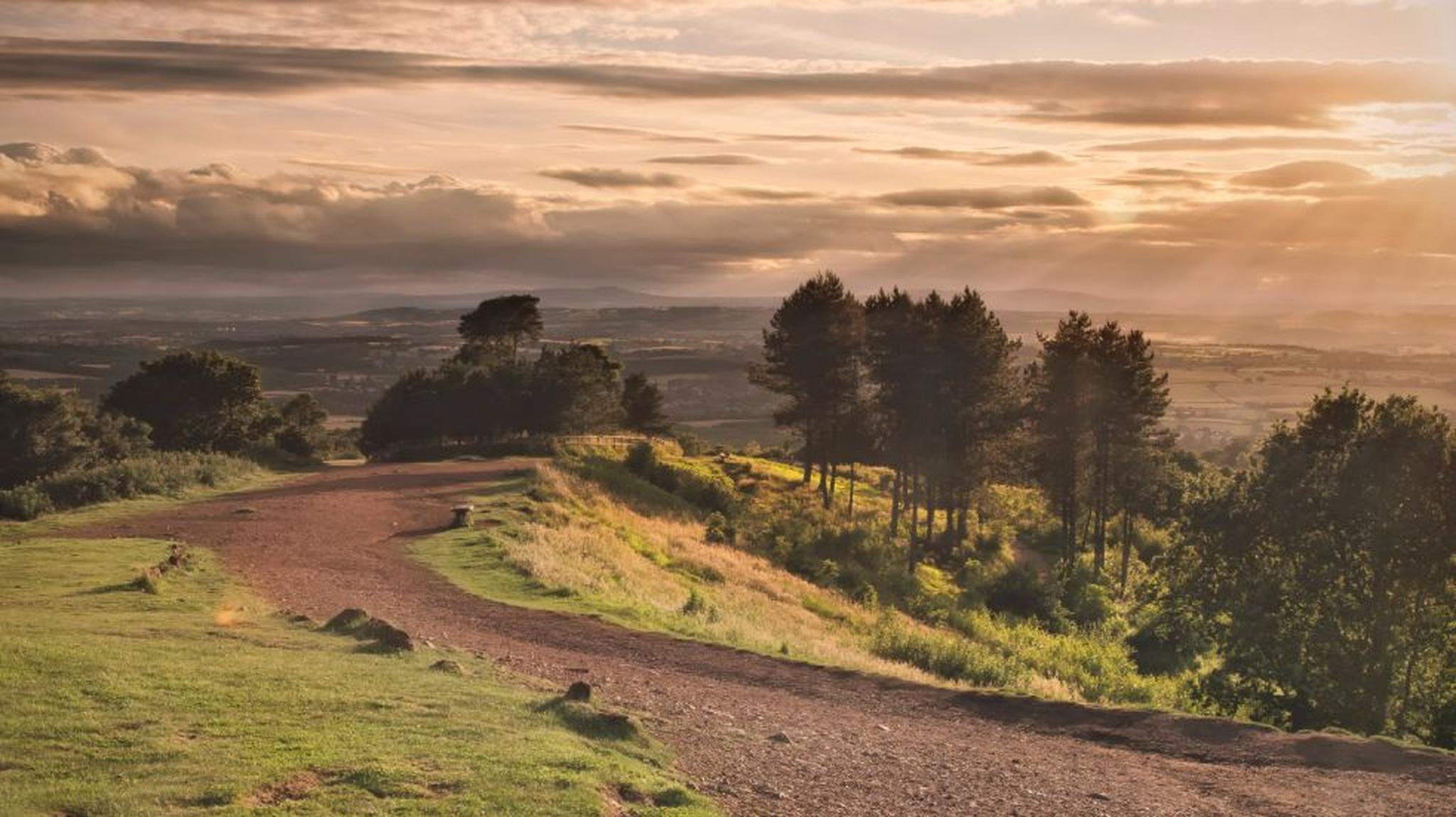 A dirt path winds through a grassy hilltop, lined with trees, under a dramatic sunset sky with sunrays and clouds.