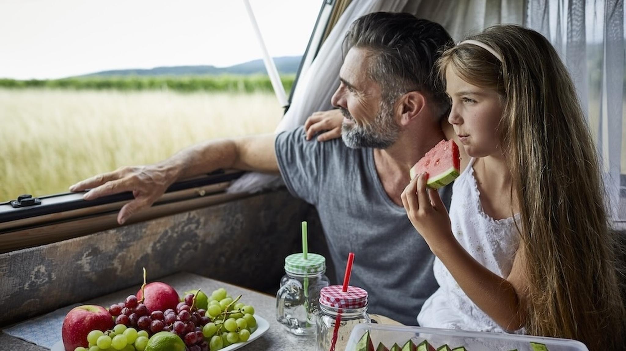 Father and daughter in a camper, enjoying watermelon and fresh fruit, gaze out at a scenic field through the window