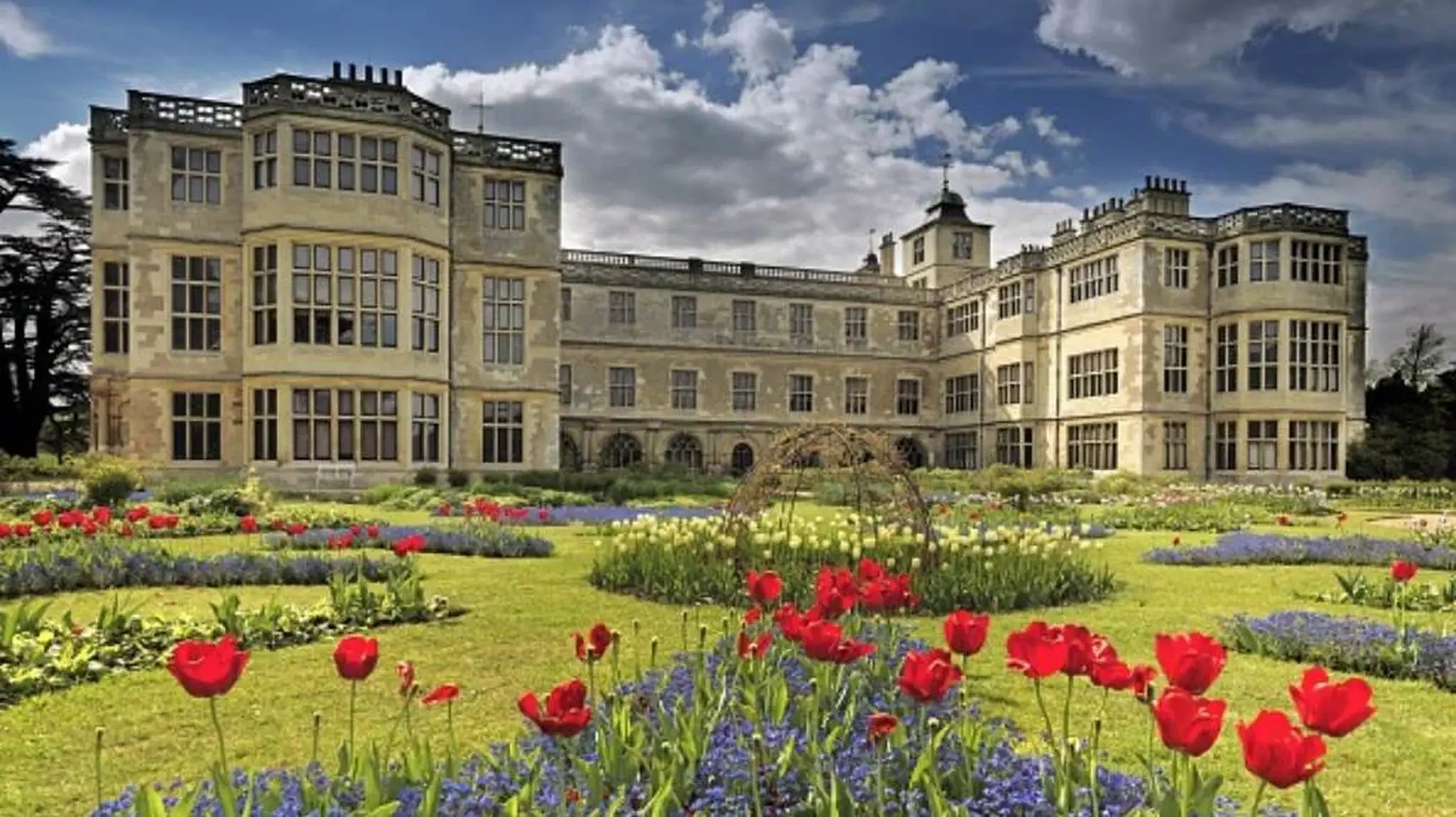 Historic mansion with ornate windows, surrounded by gardens and a vibrant mix of red tulips and blue flowers