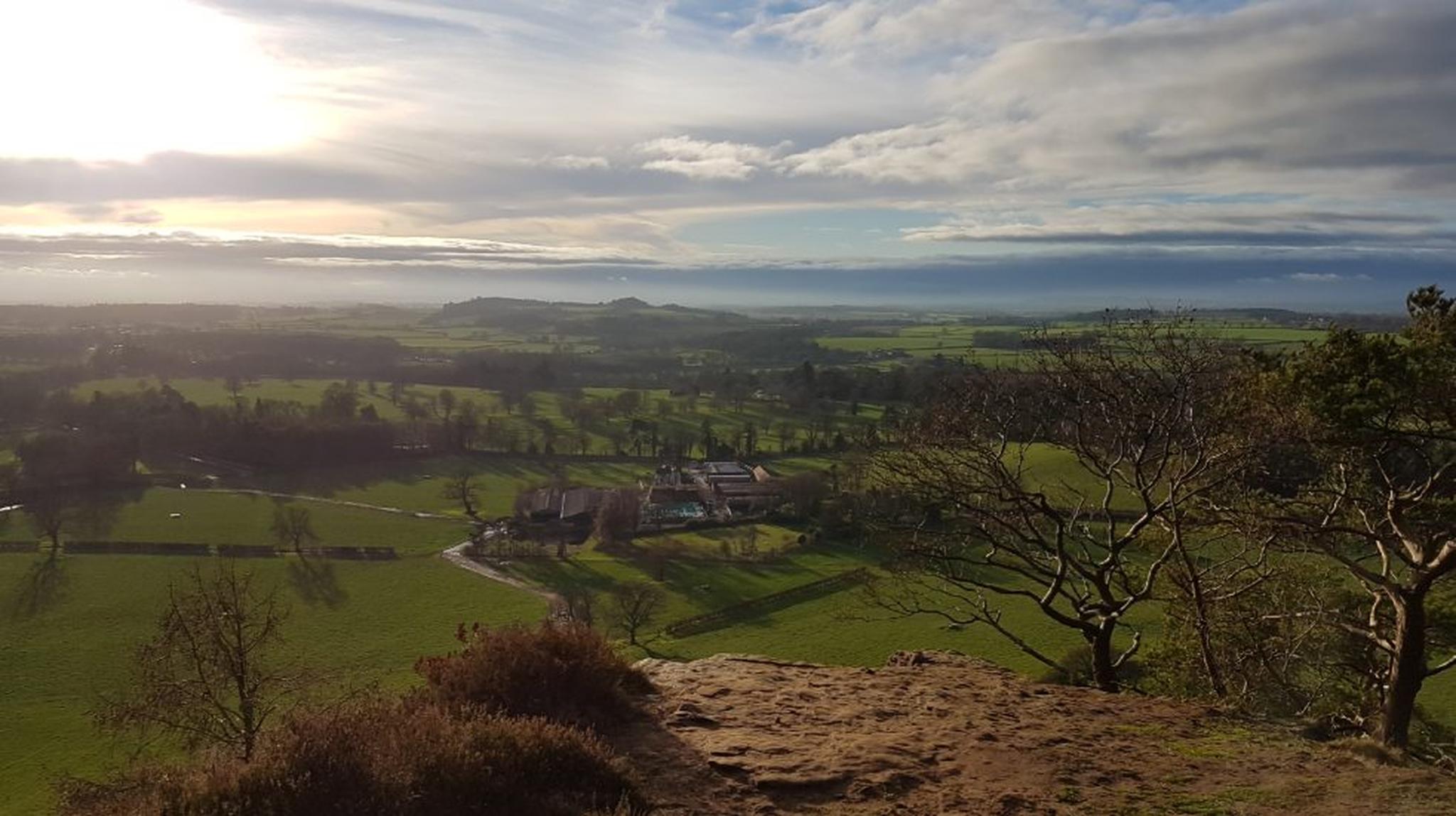 A scenic landscape from a hilltop overlooking green fields, a farmhouse, and a vast horizon under a partly cloudy sky.