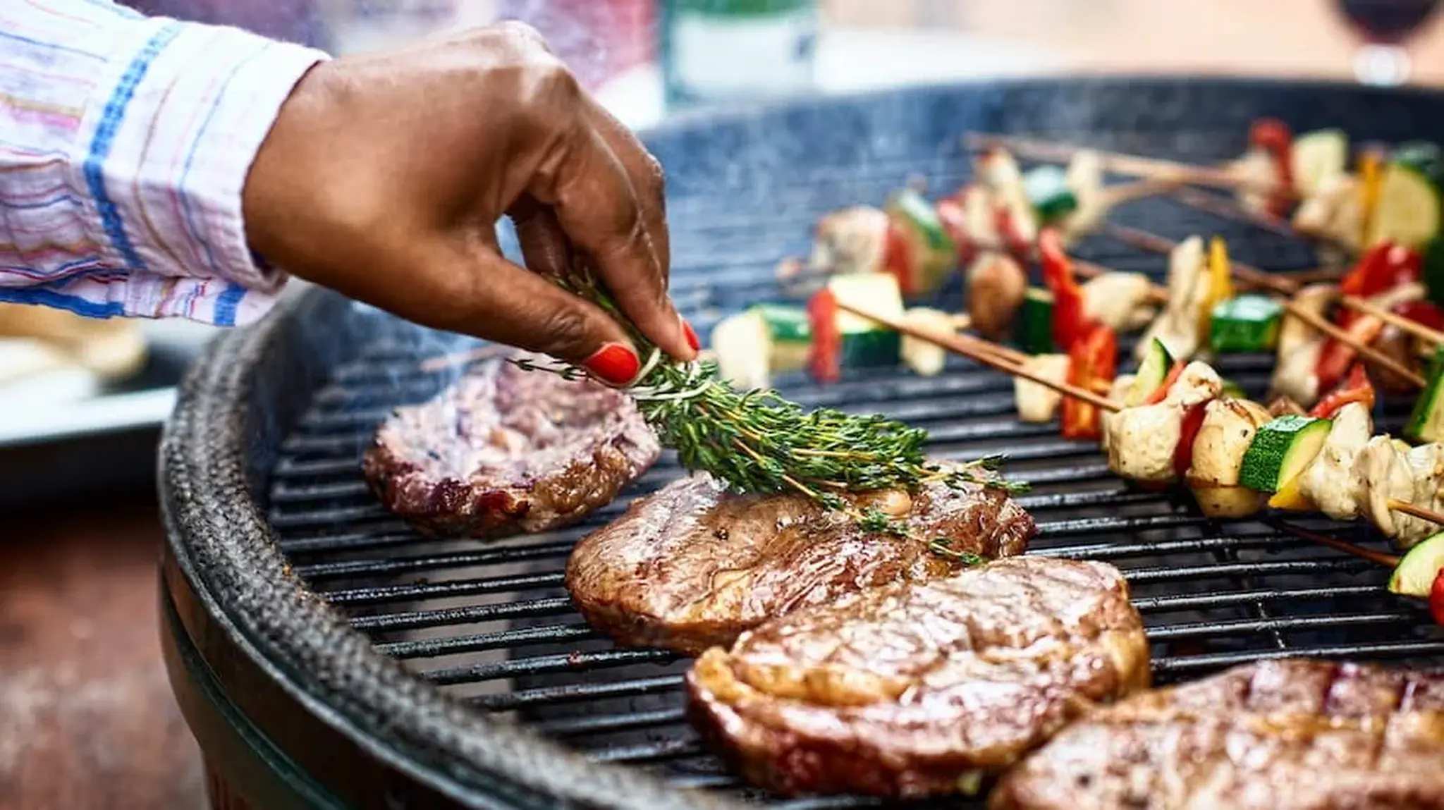 Hand seasoning steaks with herbs on a grill alongside skewers of vegetables