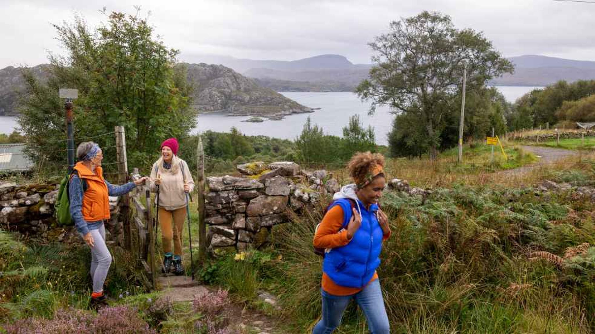 Three people hiking near a stone fence, with a scenic lake and mountains in the background on a cloudy day.