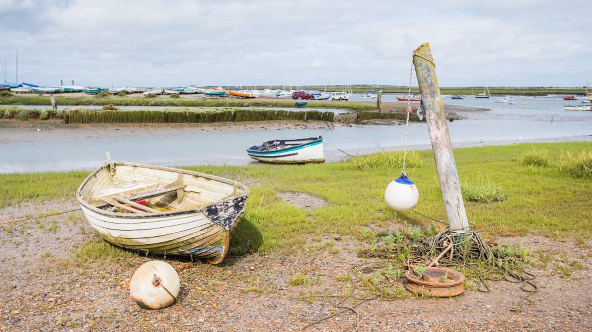 Rowboats resting on grassy shore near a calm river, with mooring post and buoy under a cloudy sky. Boats and fields visible in the background.